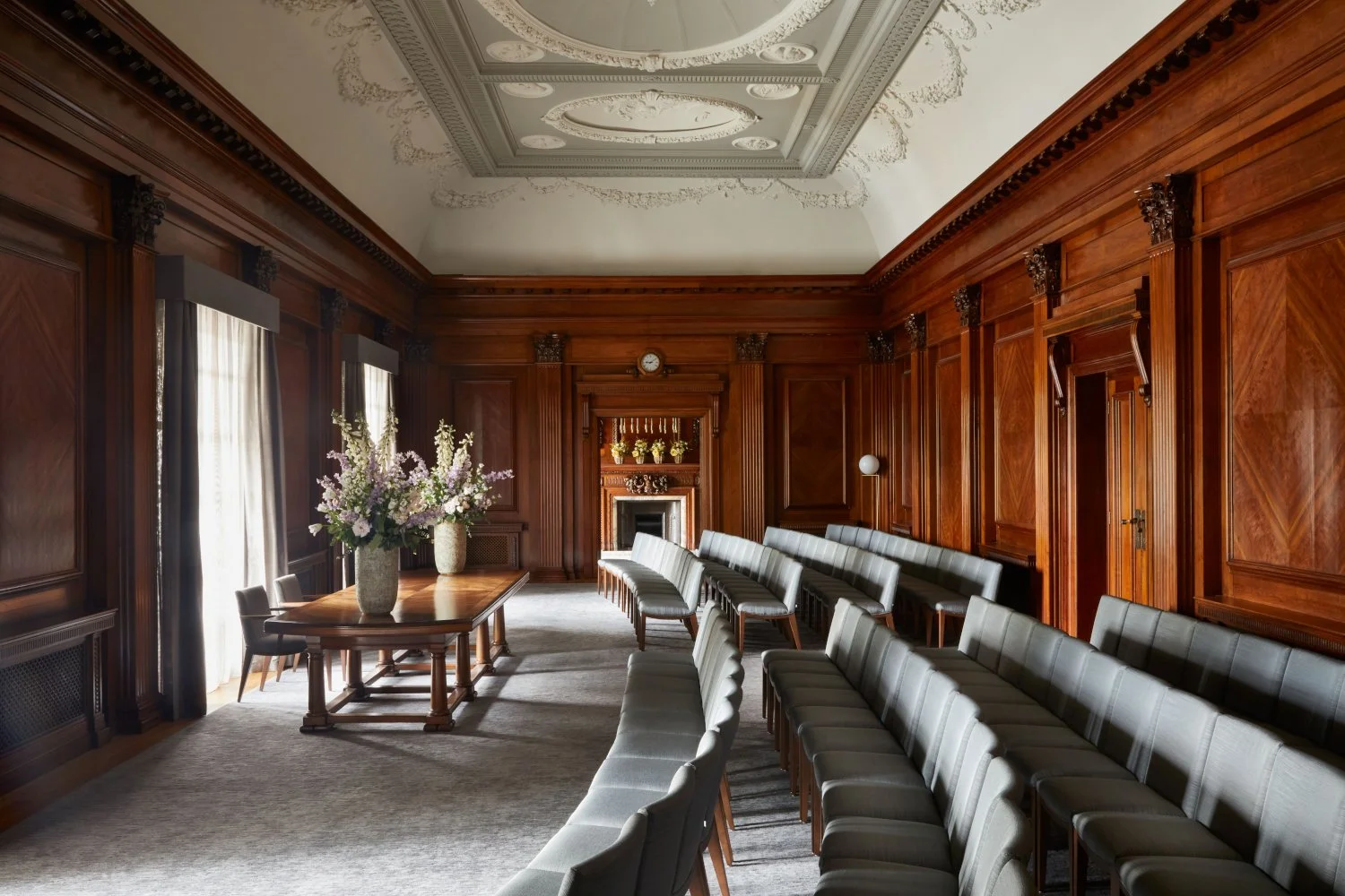 木彫りの装飾が施されたクラシックな会議室または式典会場の内装。長い木製のテーブルに花の飾り、白い椅子が並び、木の壁と窓にはカーテンが掛かっている。
