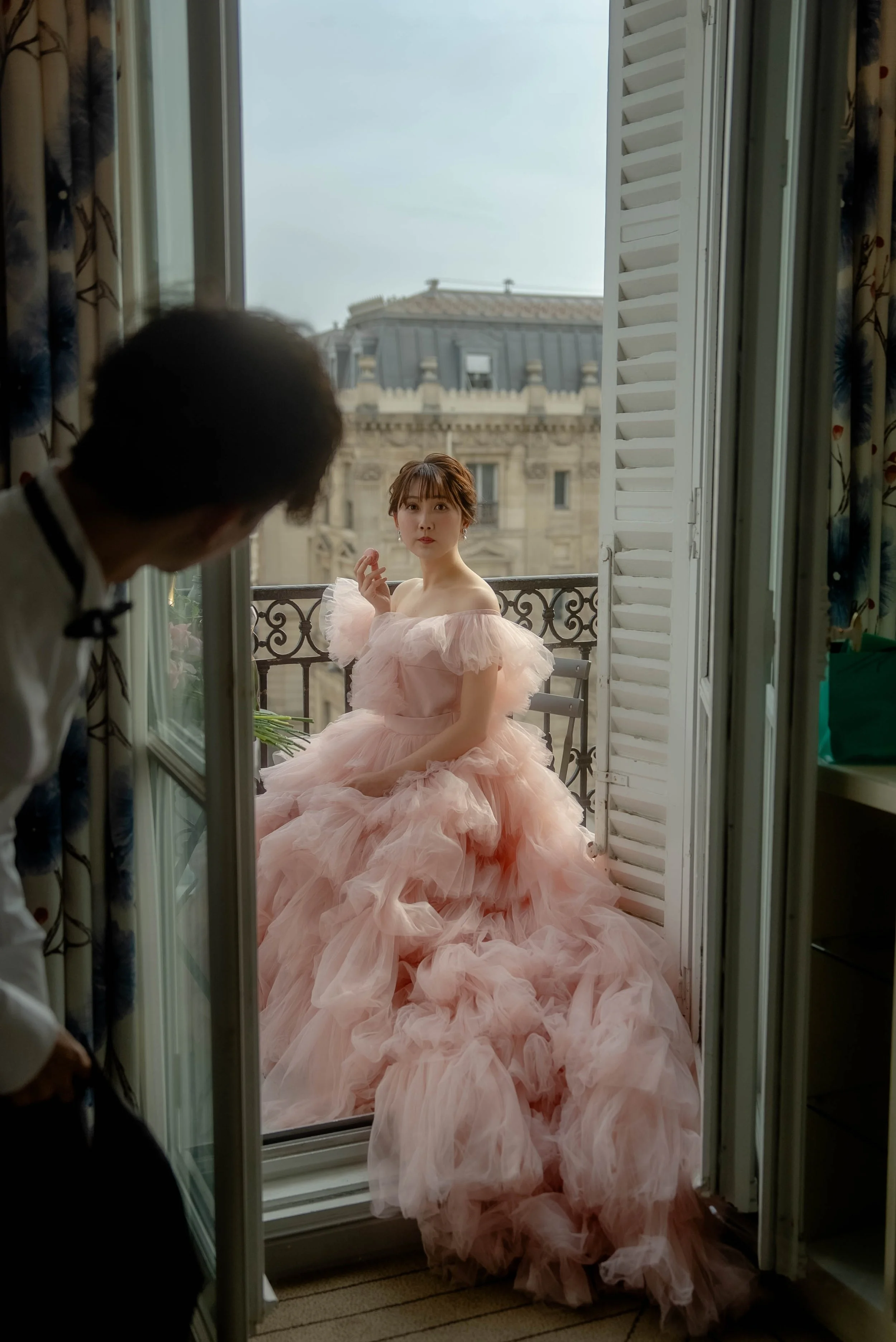 ピンクのふわふわのドレスを着た女性がバルコニーに座っている。室内にいる男性が彼女を見ている。背景には青空と建物の屋根が見える。