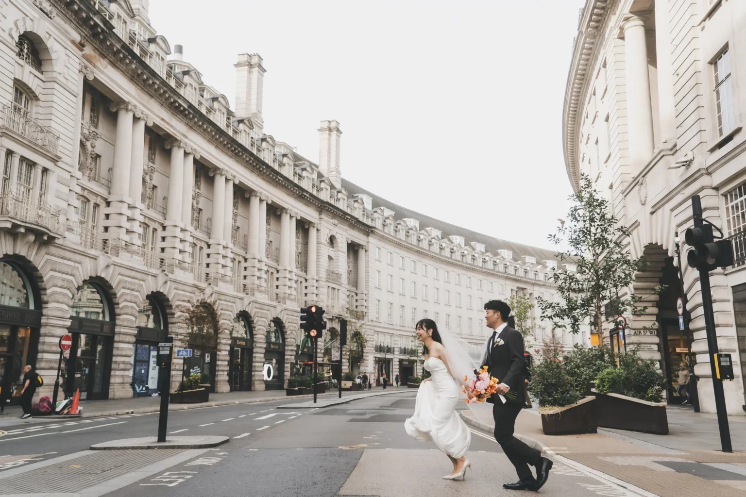 結婚式のカップルが通りを渡っている風景。女性は白いドレス、男性は黒いスーツを着ており、花束を持っている。背景には古典的な建物が円形に並んでいる都市の風景が写っている。