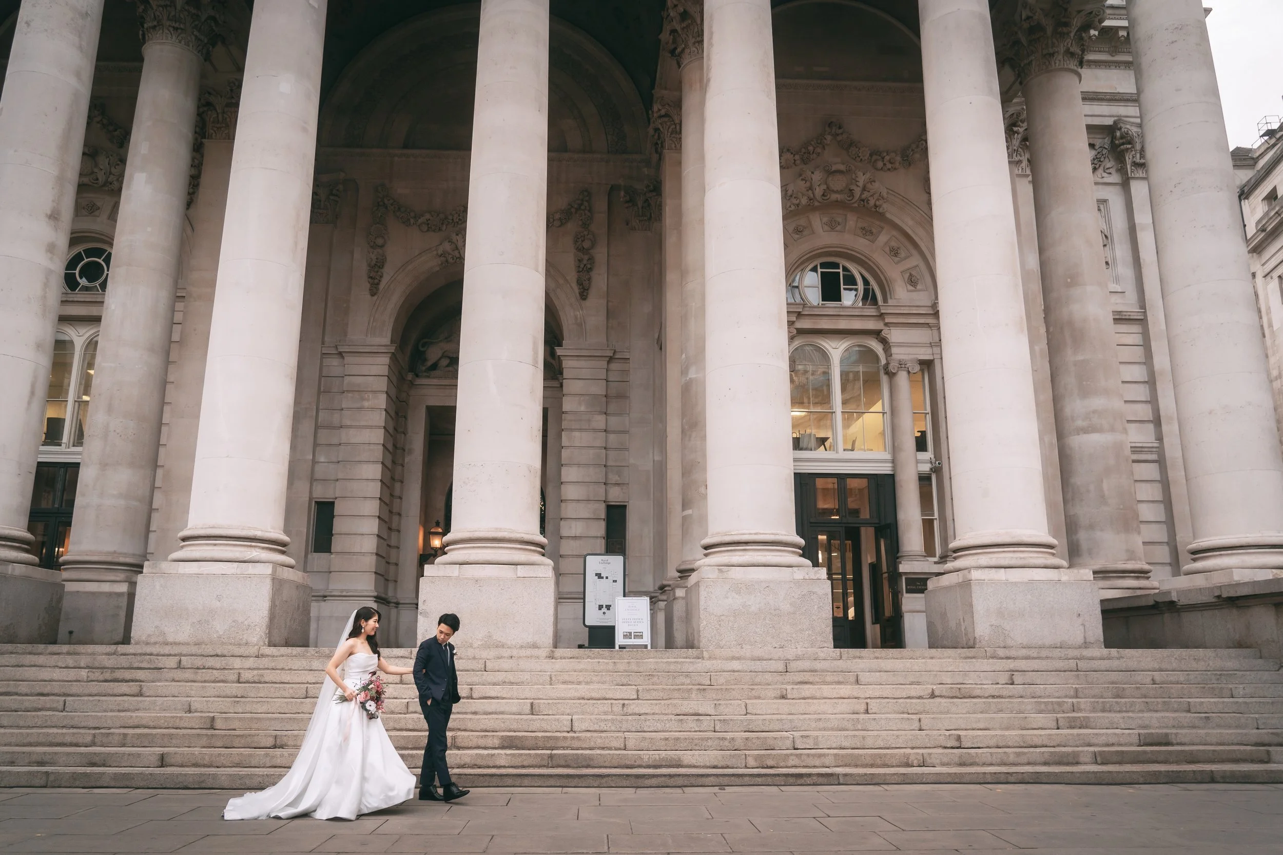 結婚式のカップルが結婚式場の階段を下りている。女性はウェディングドレスを着て、花束を持っている。男性はスーツを着ている。背景には大きな古典的な建築の柱と入り口が見える。