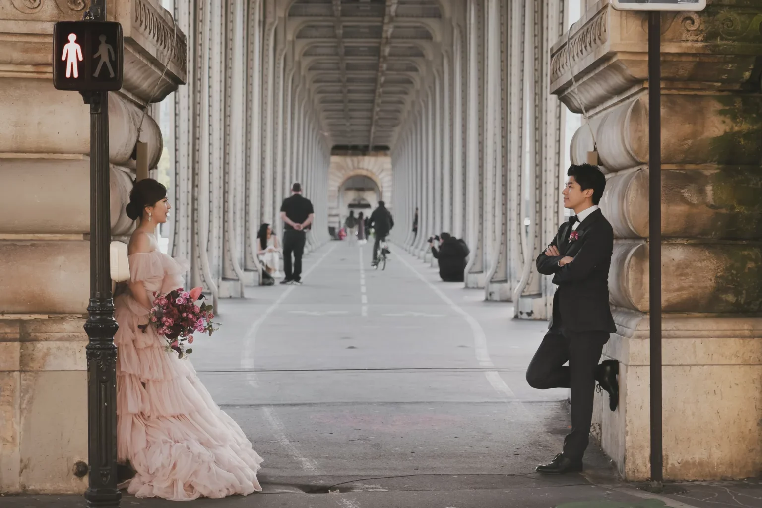 橋の下で写真を撮る結婚式のカップル。左側はピンクのドレスの花嫁、右側は黒のスーツの新郎。背景には人々と自転車がいる。