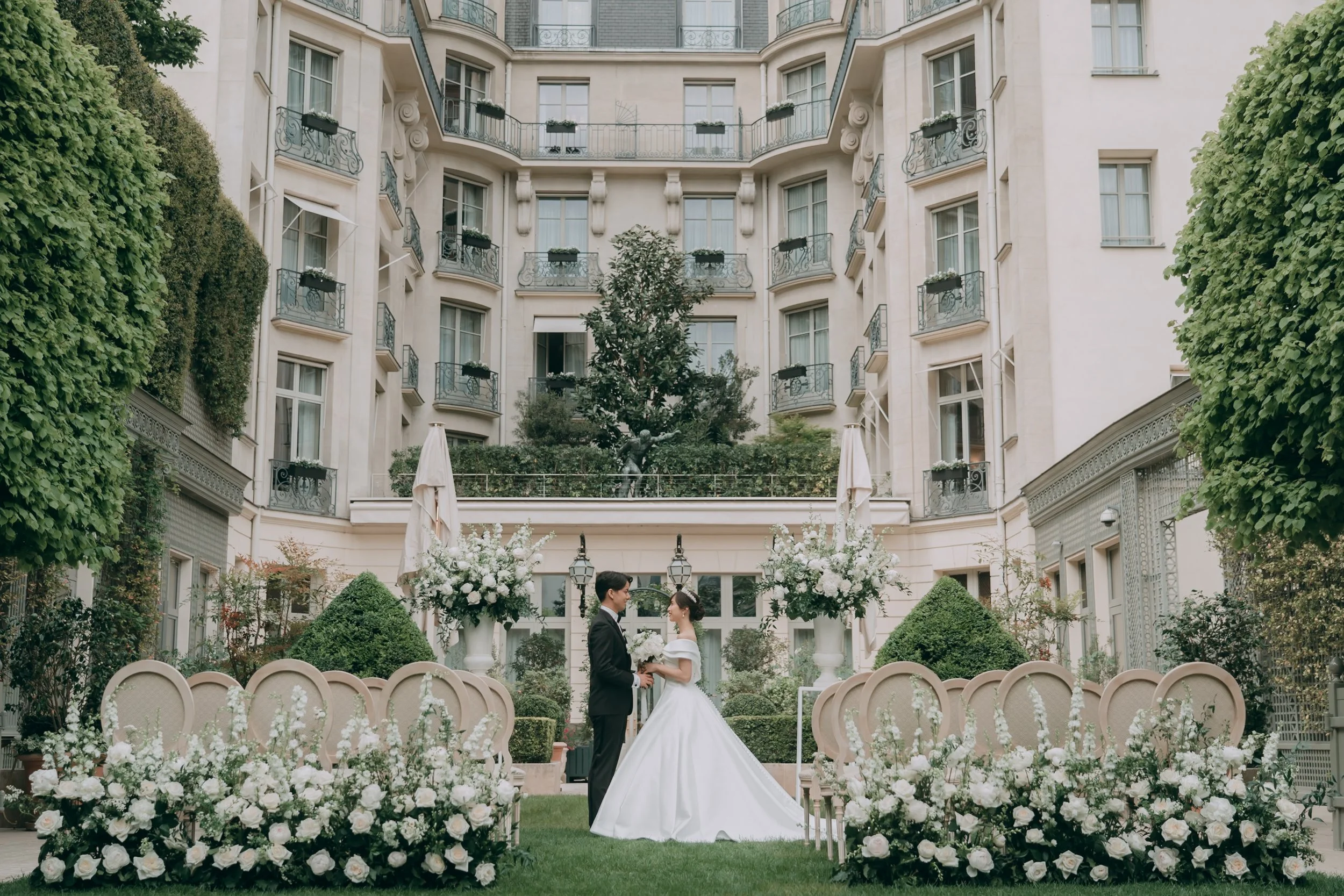 結婚式の撮影風景。新郎と新婦が庭園で向かい合っている。周囲に白い花と装飾が配置されている。背後には白い建物と家具、緑の木々。