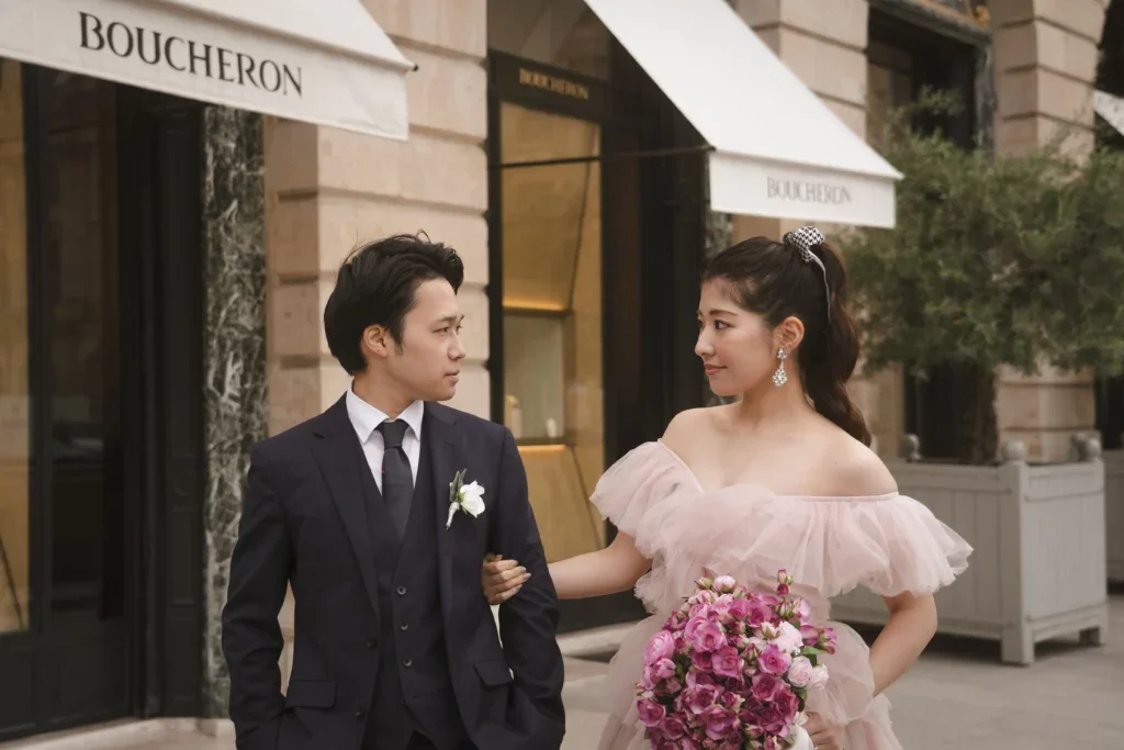 結婚式の外で新郎と新婦が互いに見つめ合う。新婦はピンクのドレスを着て花束を持ち、新郎は黒のスーツを着ている。背景にブタシェロンの看板が見える.