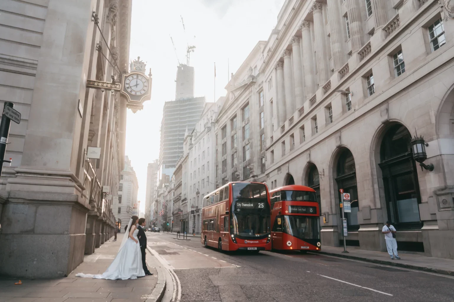 街並みの中央に赤いバスと二階建てバスが停車している。左側にウェディングドレスを着た女性と男性が通行人として歩いている。背景に古い建物と高層ビルが見える。