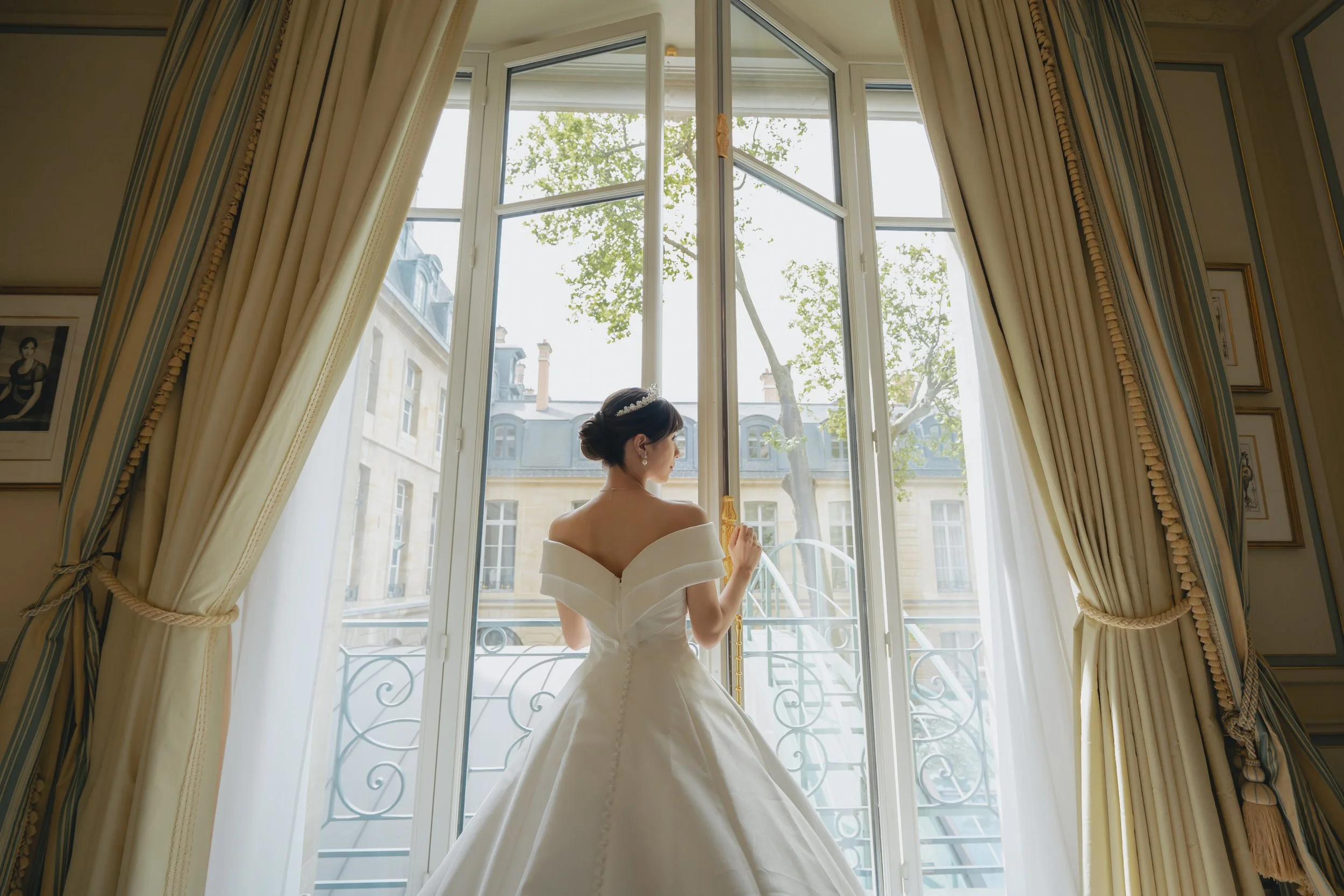 白いドレスを着た女性が大きな窓の前に立ち、外の景色を見ている。カーテンは金色で束ねられている。