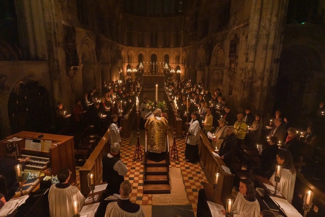 教会の内部で行われている宗教的な儀式の様子。中央に司祭の衣装を着た人物が立ち、その周りに信者や司祭の補佐をする人々が並び、蝋燭の明かりが照らしている。