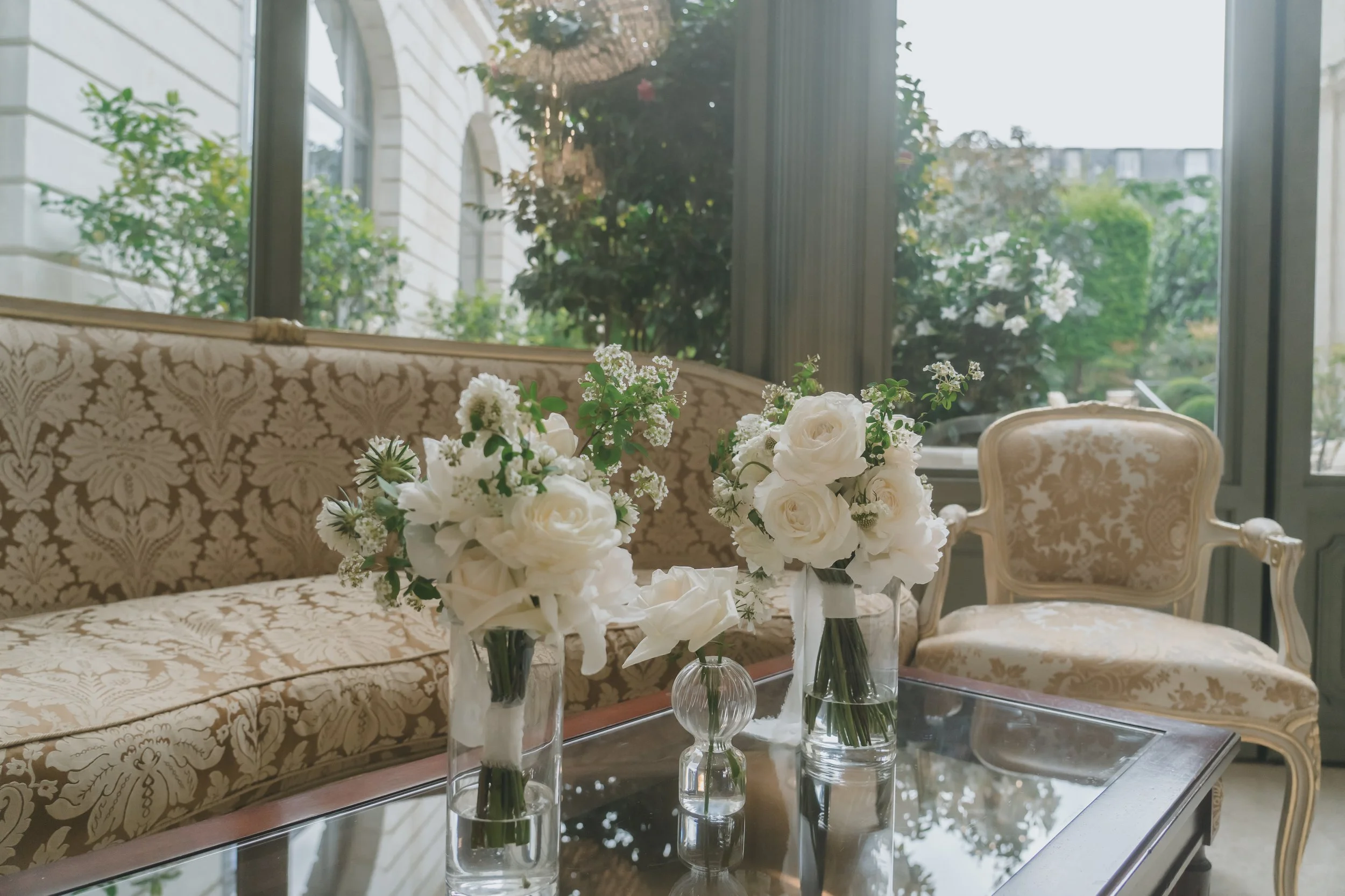 白い花のブーケを飾ったガラスの花瓶3つと、豪華な装飾が施されたソファと椅子のある室内の風景。