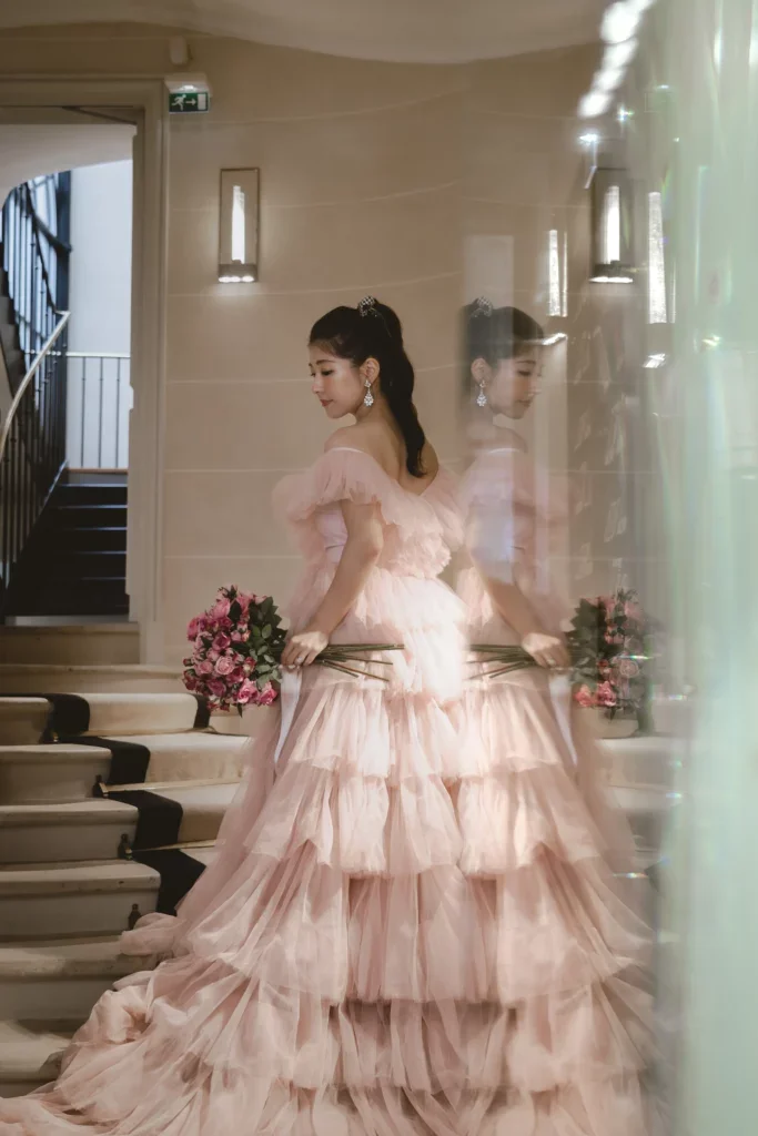 ピンクのドレスを着た女性が花束を持ち、鏡の前でポーズをとっている。