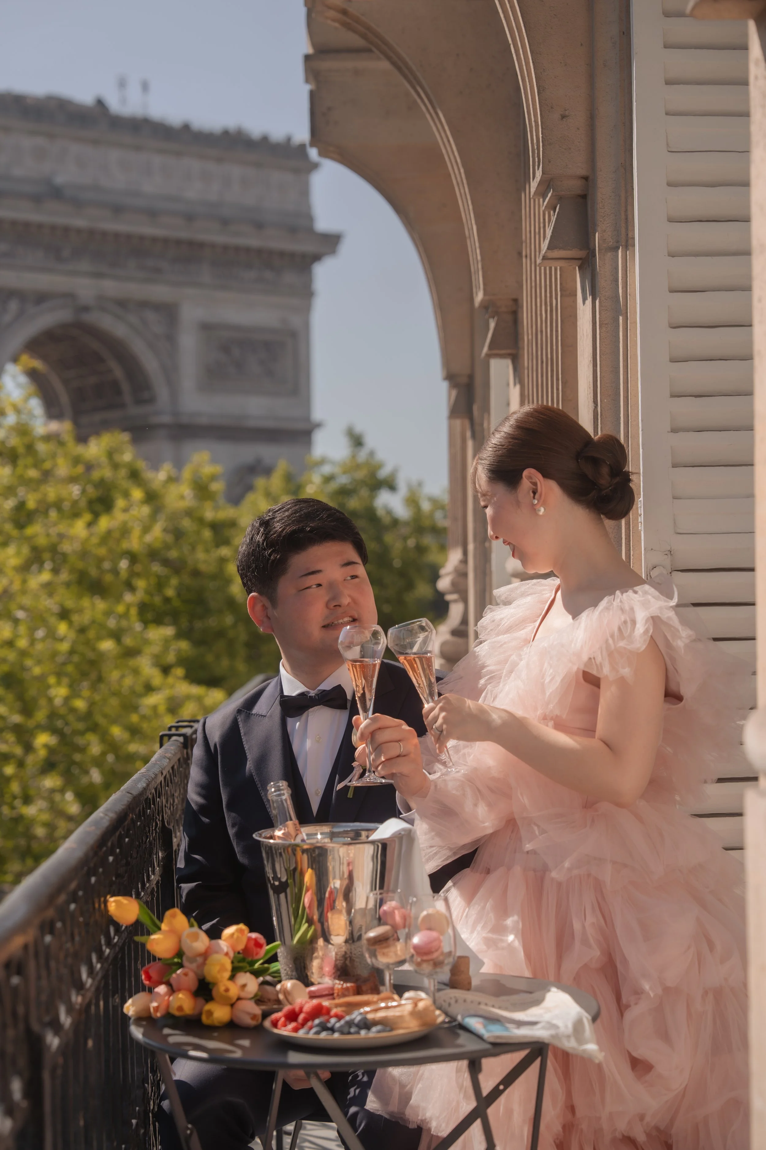 パリのエッフェル塔が背景のベランダで、花嫁と花婿がシャンパングラスで乾杯しているカップルの写真。花嫁はピンクのドレス、花婿は黒のタキシードを着ている。テーブルには花束とデザートが置かれている。