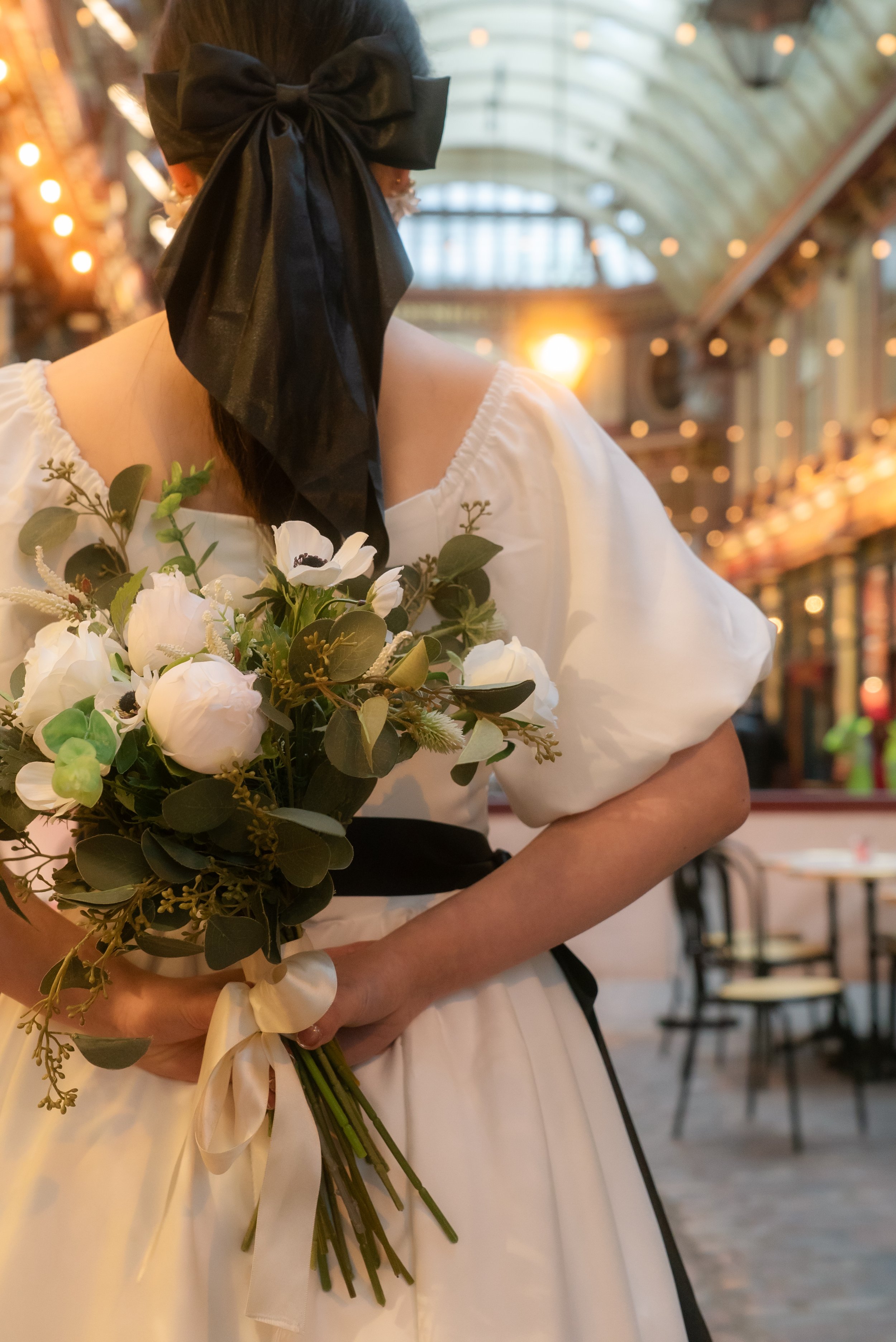 白いドレスを着た女性が花束を持ち、キャナルシティ博多の屋内市場のような場所にいます。彼女の顔は見えません。背景には暖色の照明とガラスの天井があります。