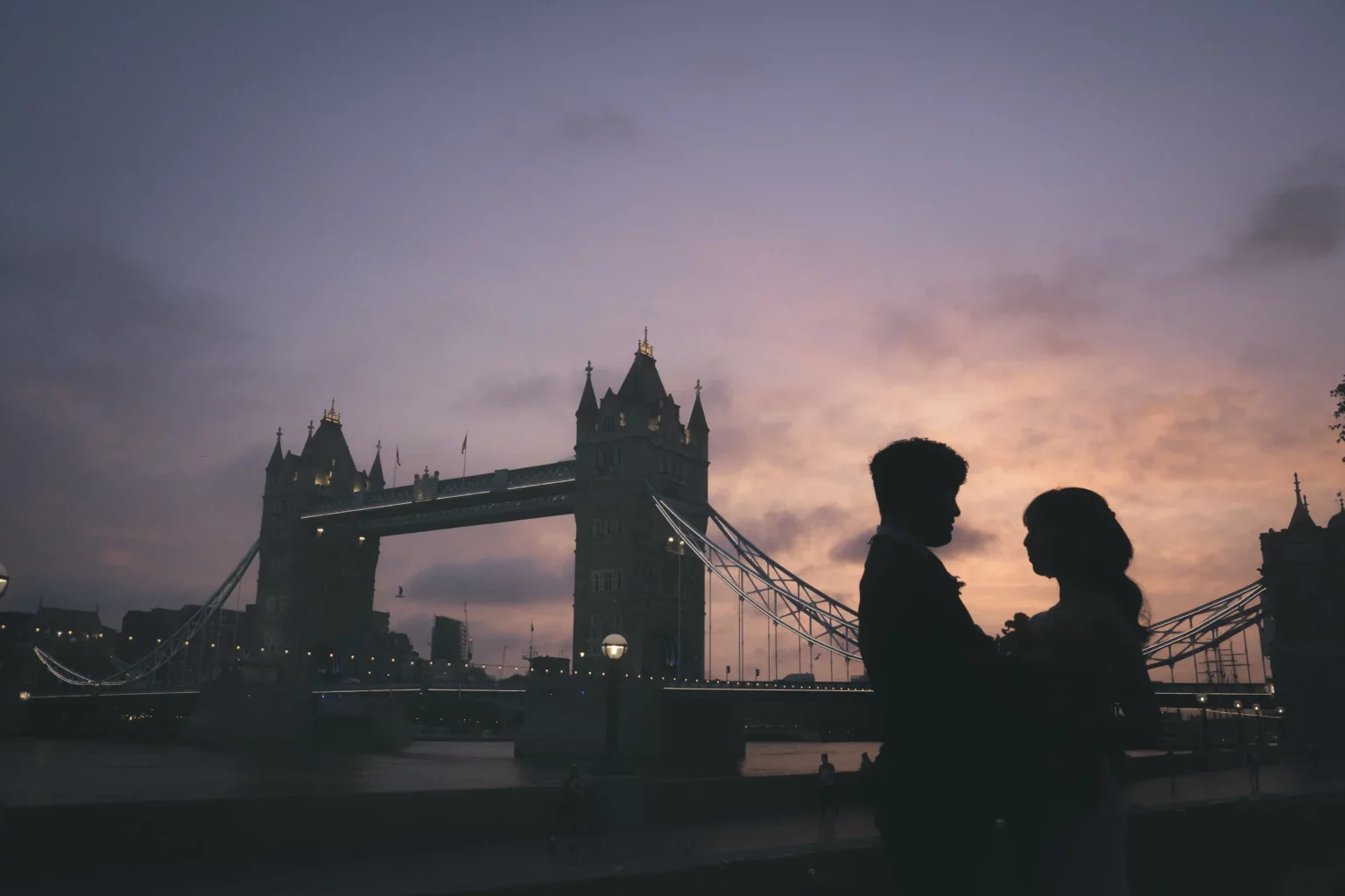 夕暮れ時のロンドンの橋の前で、カップルが手を取り合っているシルエットの写真