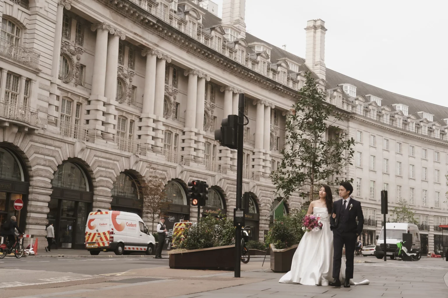 結婚式を挙げる新郎新婦が街頭を歩いている。女性は白いドレスと花束を持ち、男性はスーツを着ている。背景に古典的な建物と交通信号機が見える。