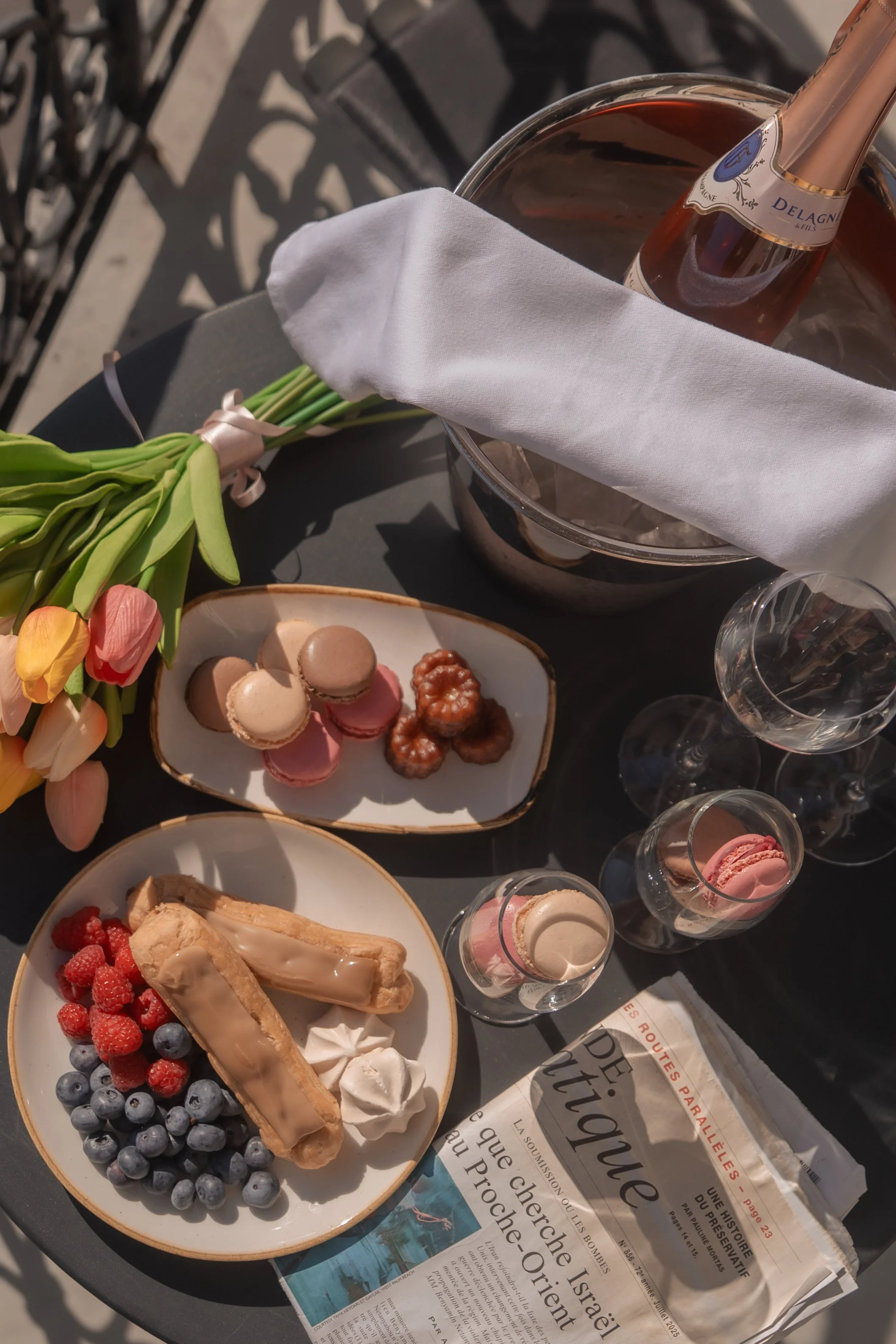 満開のチューリップの花束、トレーにのったマカロンやフルーツのケーキ、シャンパングラスと瓶、新聞紙が置かれたテーブル上のセッティングです。