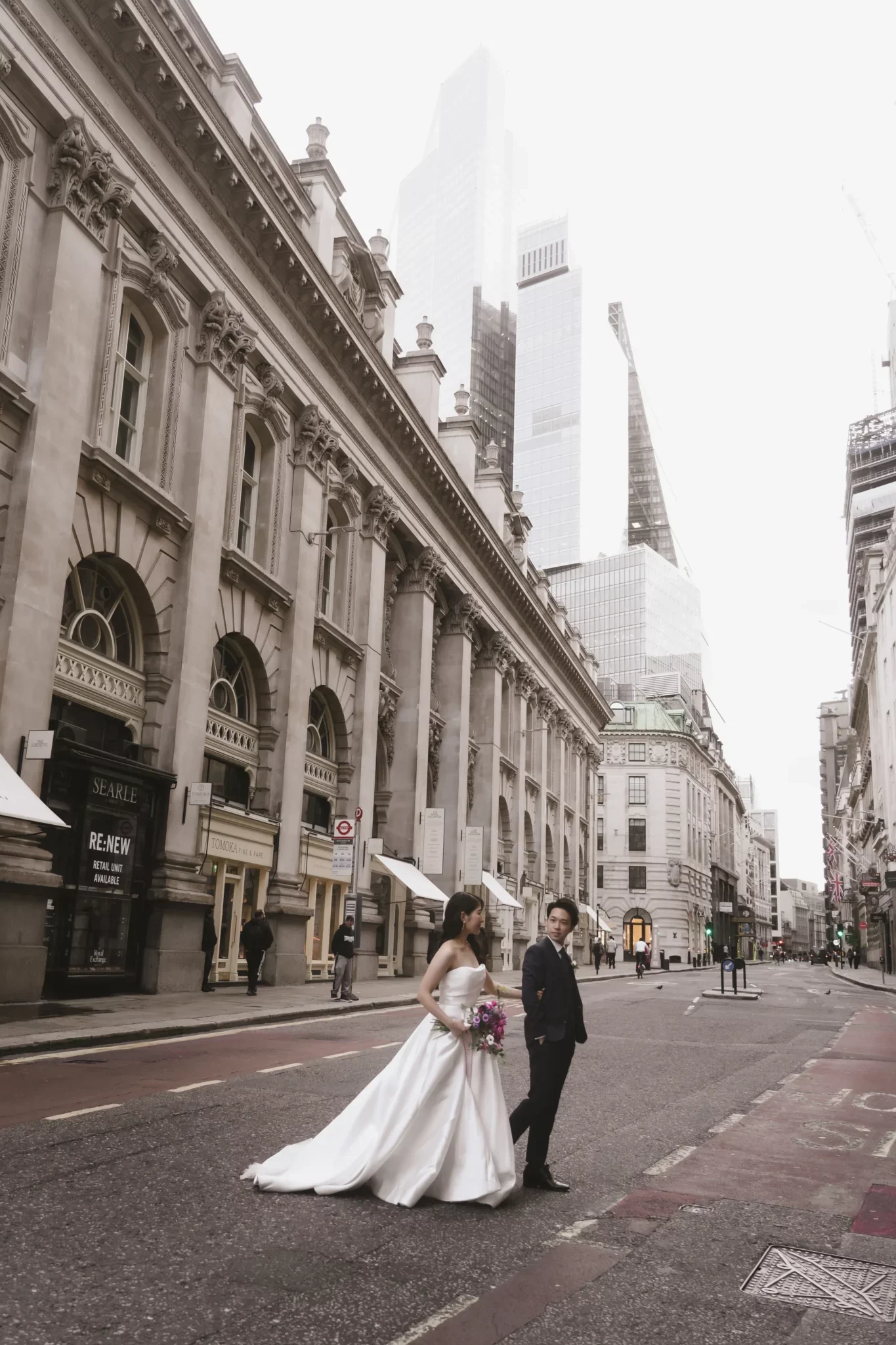 結婚式をしたカップルが都会の通りに立っている風景。女性は白いドレスを着て花束を持ち、男性はスーツ姿。背景には歴史的な建物と現代的な高層ビルが見える