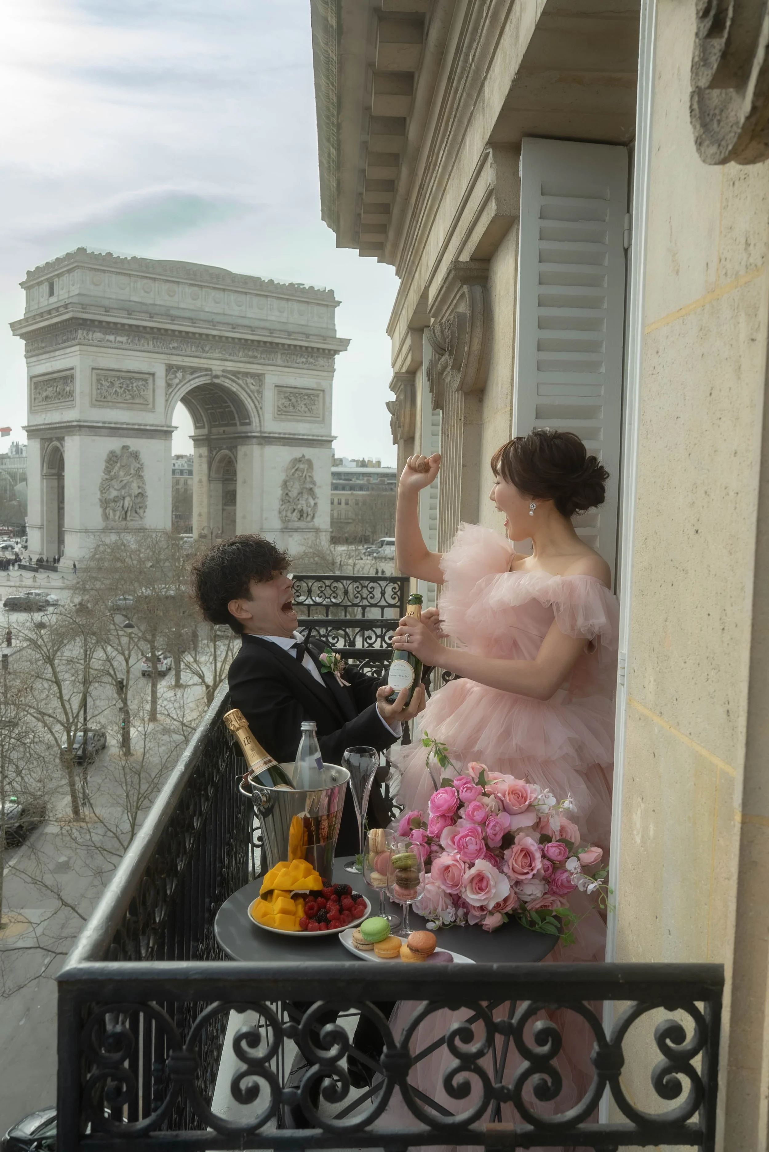 パリのエッフェル塔を背景に、祝福のシーンで女性が腕を上げて喜び、男性がプロポーズしている、バルコニーの上の結婚式の写真。テーブルにはシャンパン、ケーキ、フルーツ、花束が置かれている.