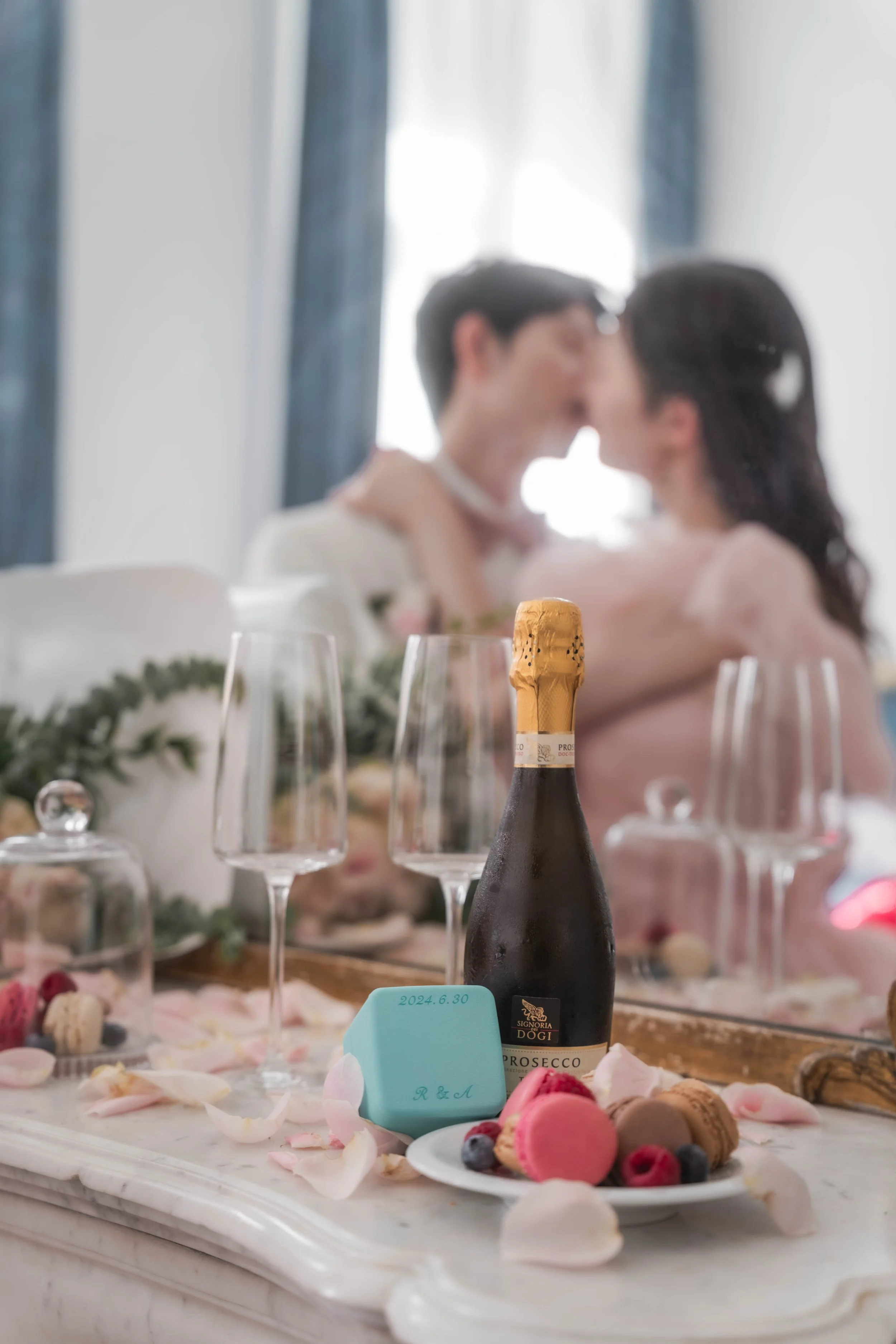 結婚式のカップルで乾杯の前に飾られたテーブル。シャンパン、マカロン、 berries、花びらなどのデザートと装飾。背景に新郎新婦がキスしているシルエットがぼやけて映る。