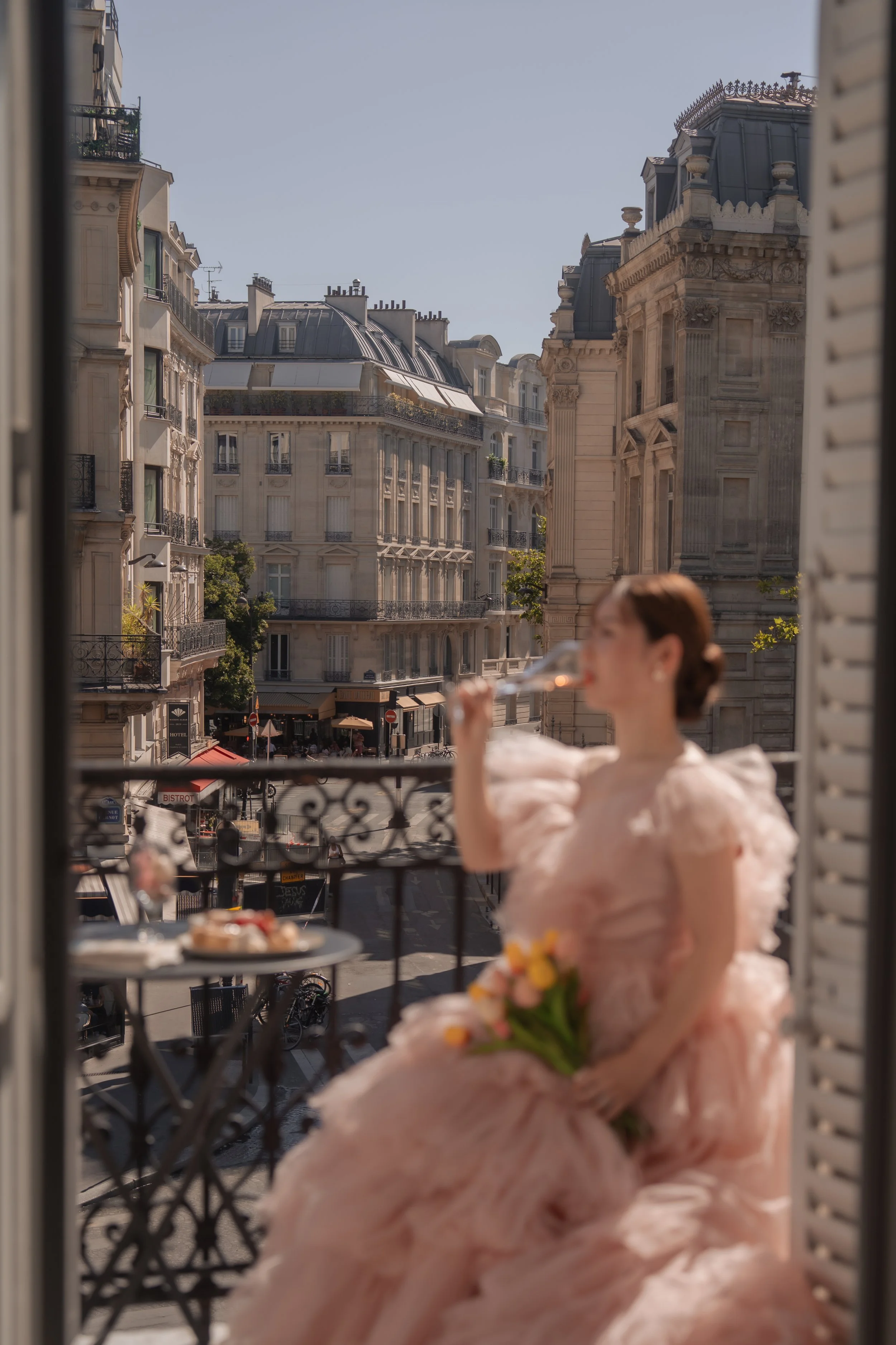 パリの都市景観を背景に、ピンク色のドレスを着た女性がバルコニーでシャンパンを飲みながら花束を持つ