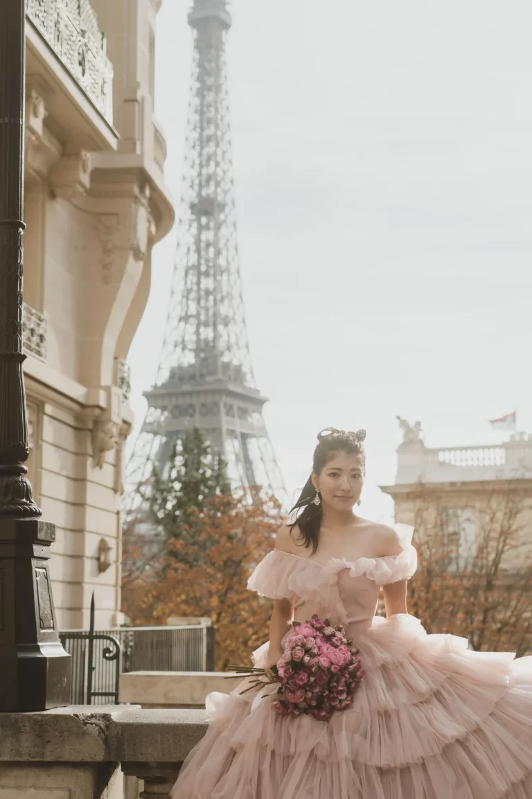 パリのエッフェル塔の側で、ピンクのドレスを着た若い女性が花束を持って微笑んでいる。