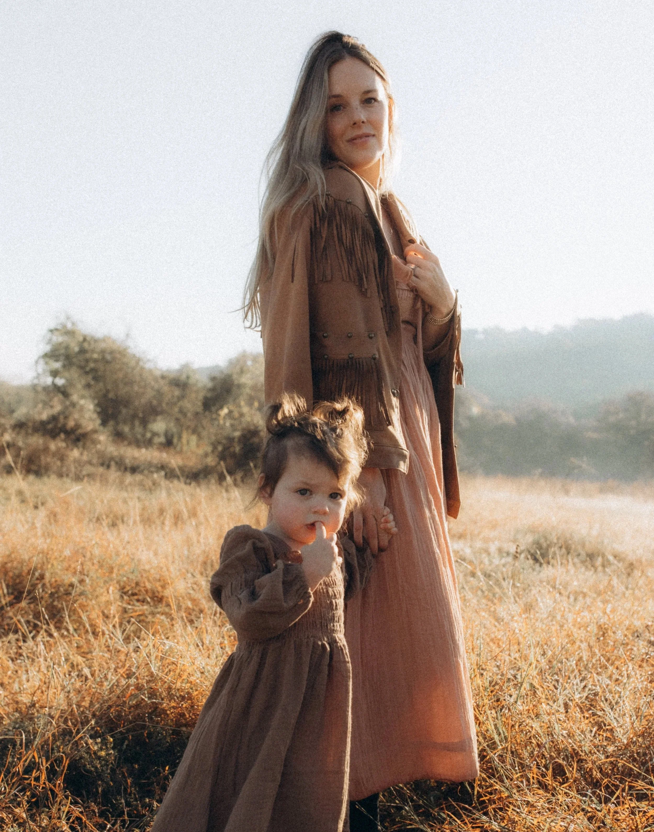 A woman and a young girl standing hand in hand in a field during daytime with clear sky.