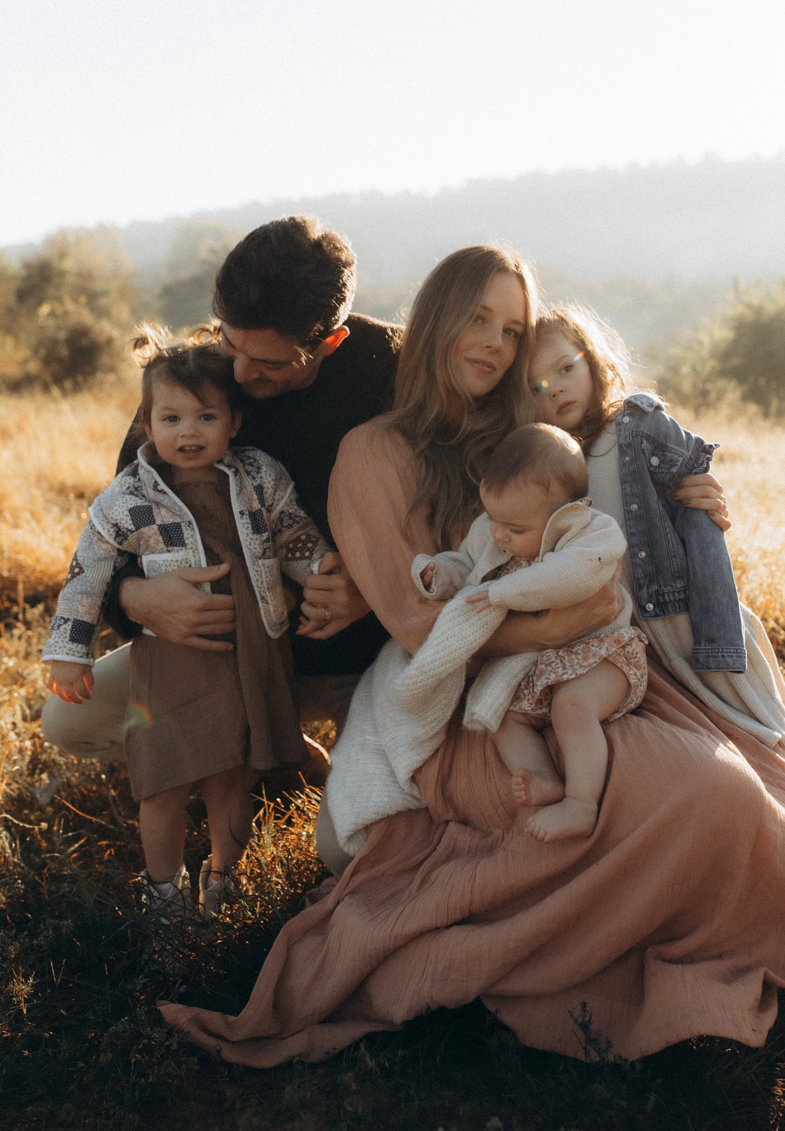 Family of five outdoors in a grassy field during golden hour, with hills in the background. The mother sits holding a toddler, the father crouches beside two young girls, one standing and the other cradled in her lap.