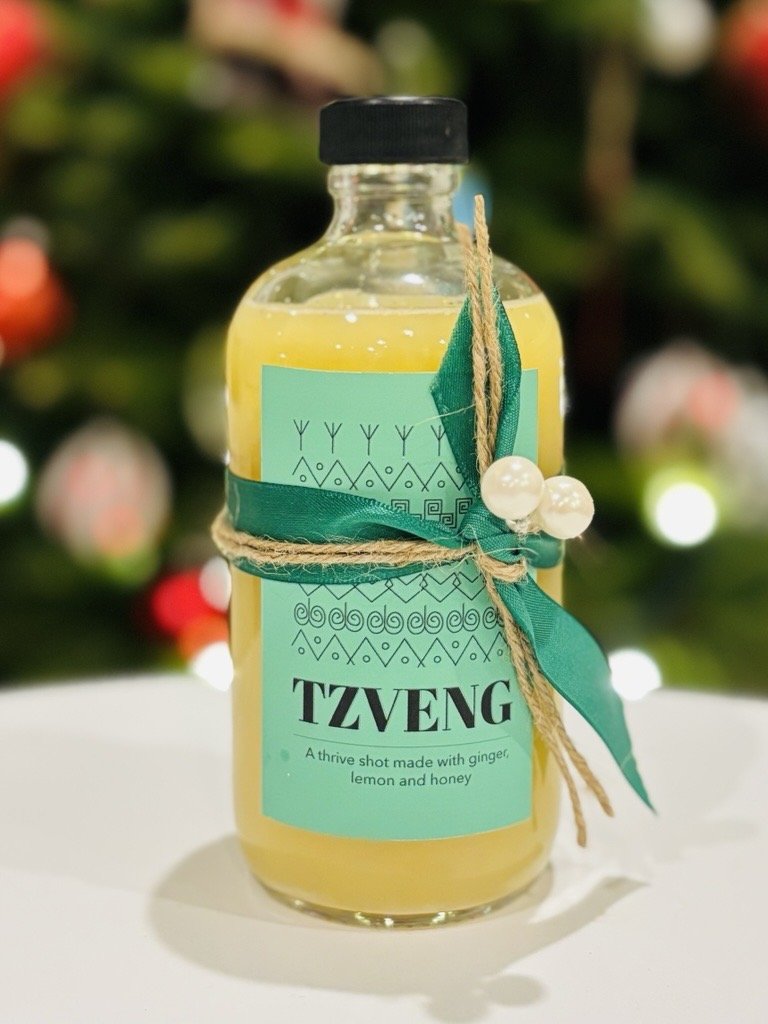 A glass bottle filled with the Tzveng ginger shot, decorated with a green ribbon, a beige twine, and white beads, labeled 'TZVENG' with a description 'A thrive shot made with ginger, lemon, and honey'.