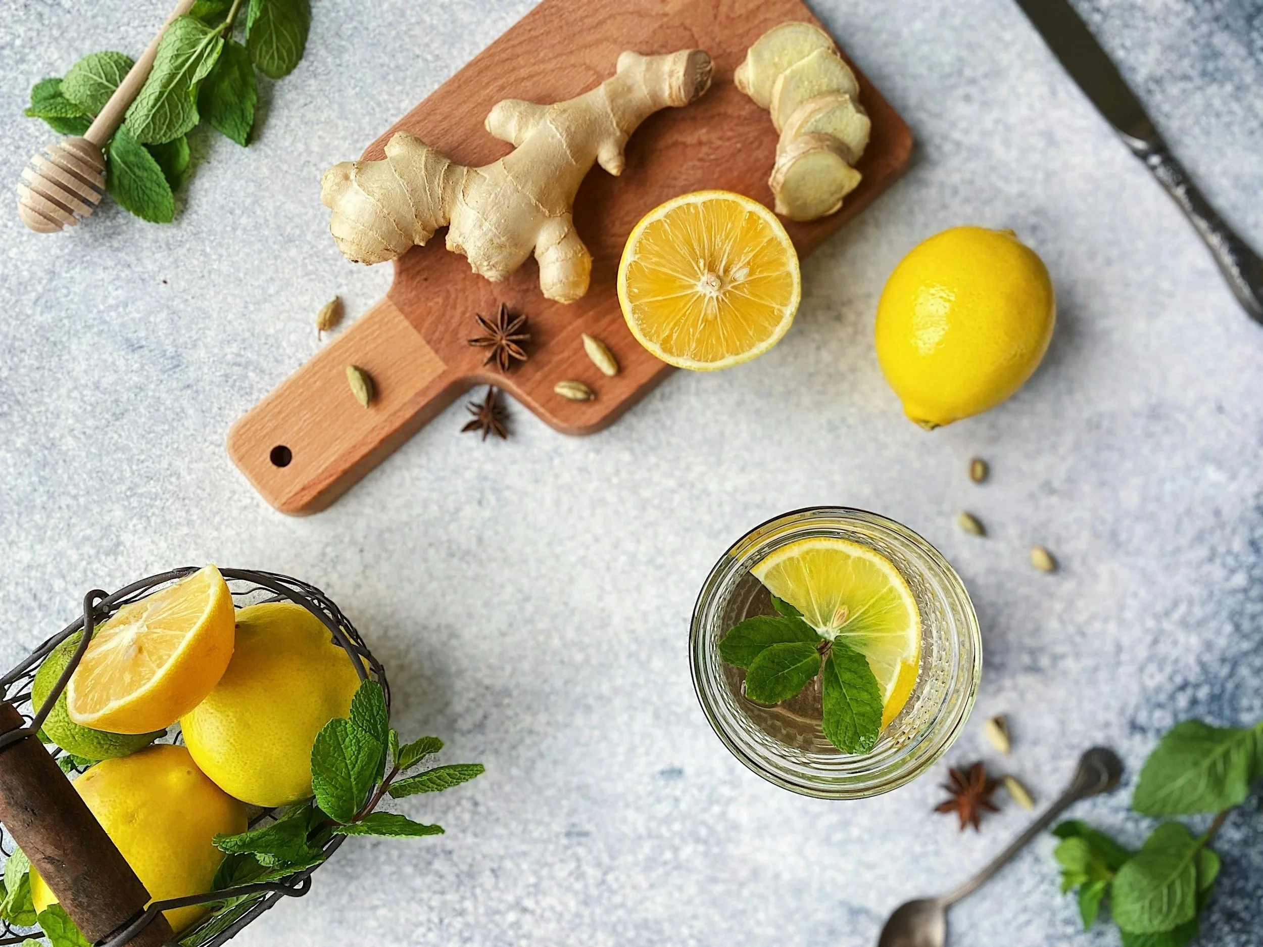 Lemon slices and ginger on a wooden cutting board, star anise and cardamom pods, fresh lemons, mint leaves in a glass jar, a glass of lemon water with mint, a honey dipper, and scattered cardamom pods on a light blue surface.