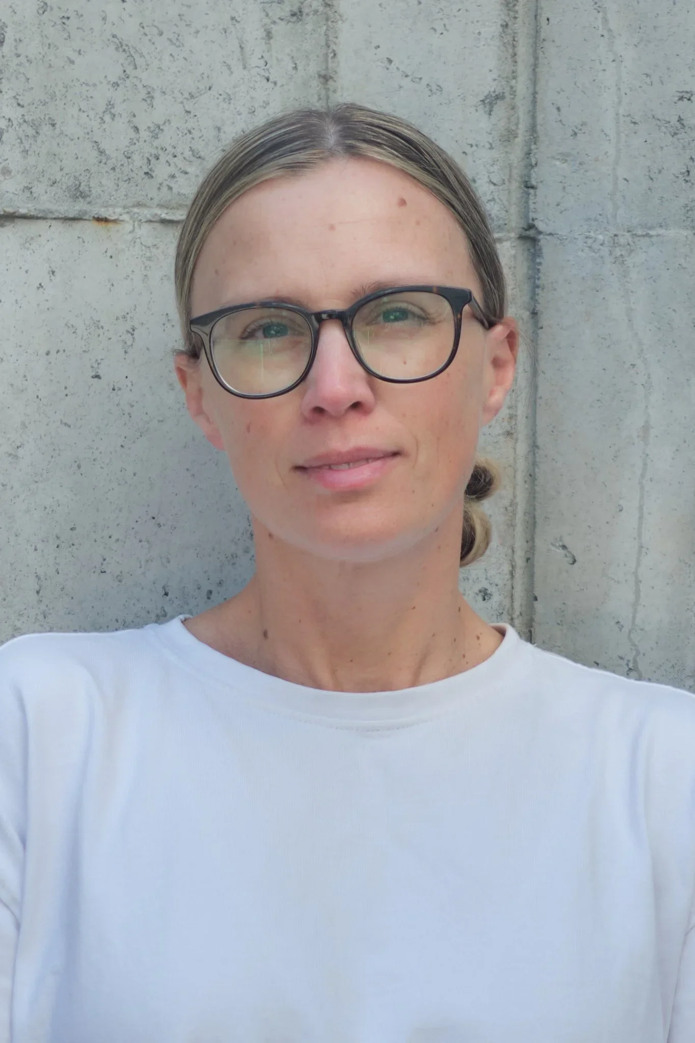 A woman with glasses and blonde hair tied back, wearing a white shirt, standing in front of a gray concrete wall.