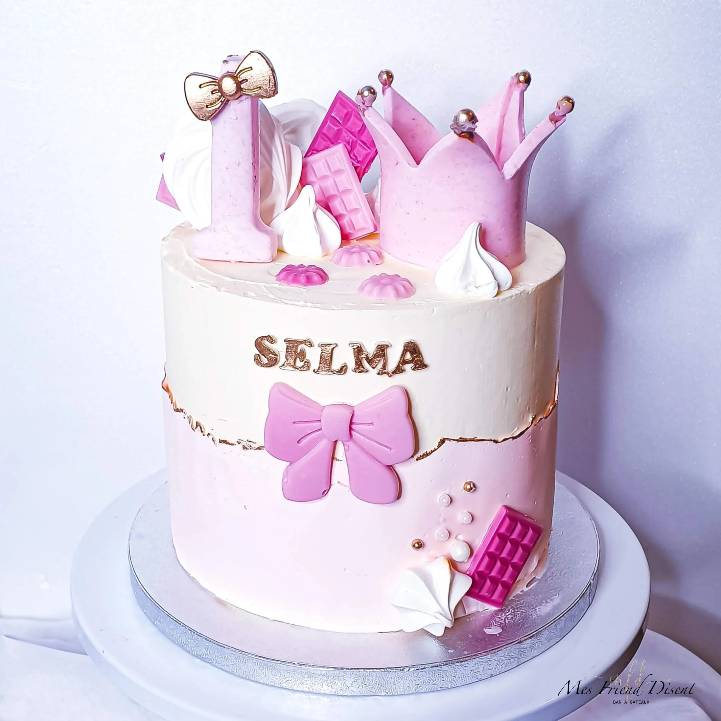 Gâteau d'anniversaire blanc et rose thème princesse, avec le nom 'Selma' écrit dessus, décoré de rubans en pâte à sucre, de morceaux de chocolat, et d'un petit papillon. Mes Friends Disent partenaire NappyLife pour la confection de vos gâteaux.