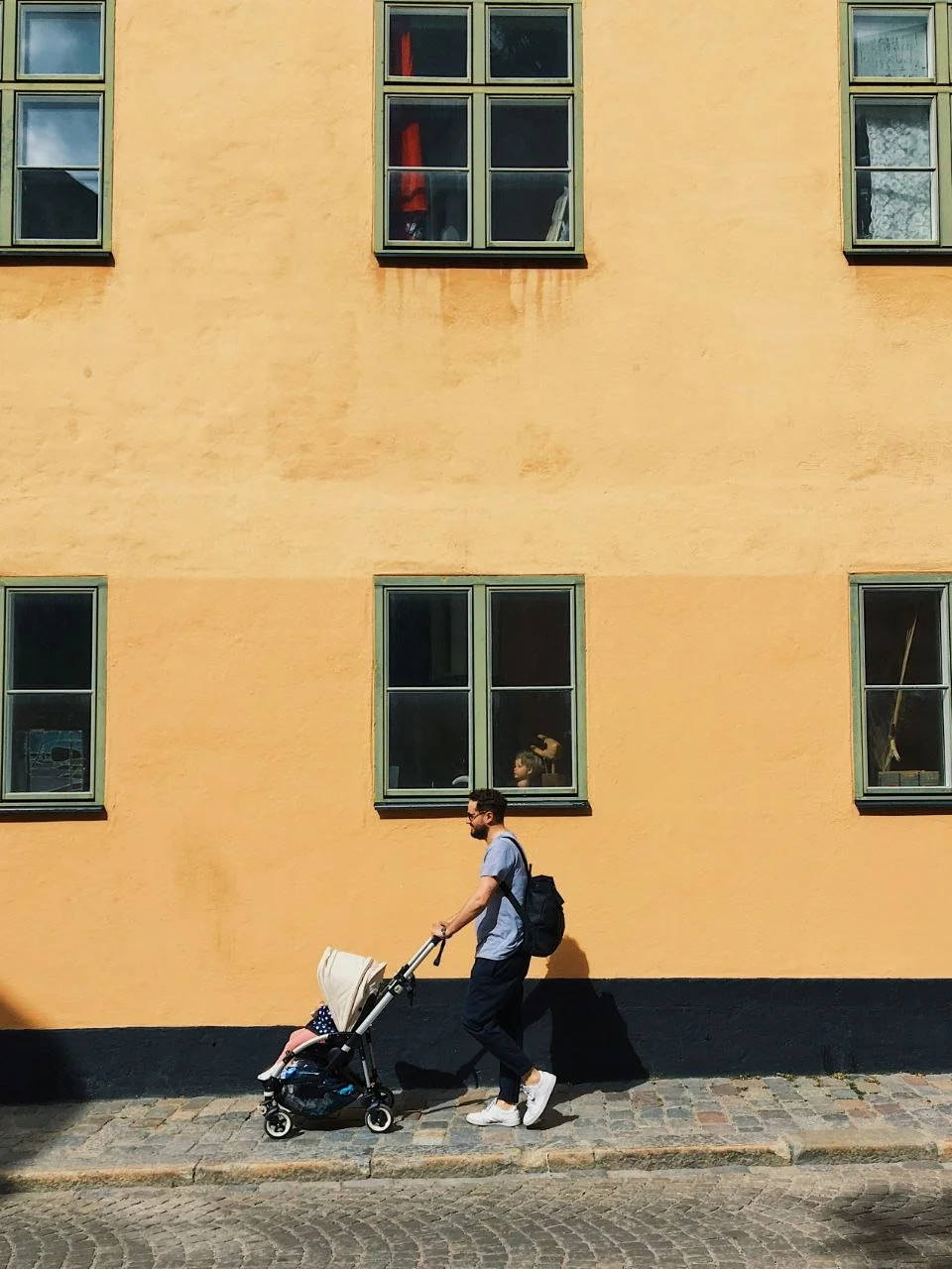Un homme poussant une poussette avec un enfant dans la rue, devant un bâtiment jaune avec plusieurs fenêtres.