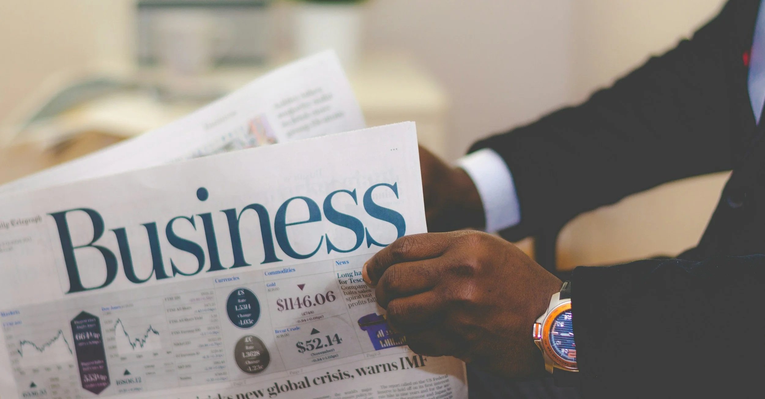 Person in a dark suit and colorful watch reading a newspaper titled 'Business' with financial and stock market information.