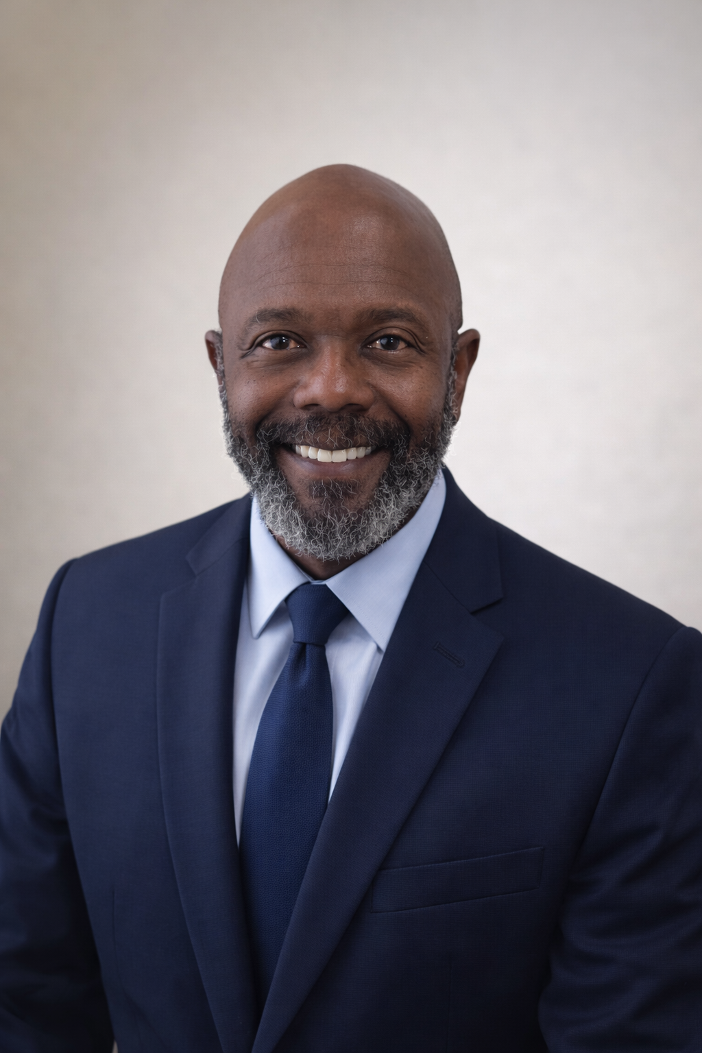 A smiling African American man in a dark blue suit, white shirt, and blue tie, looking directly at the camera against a neutral background.