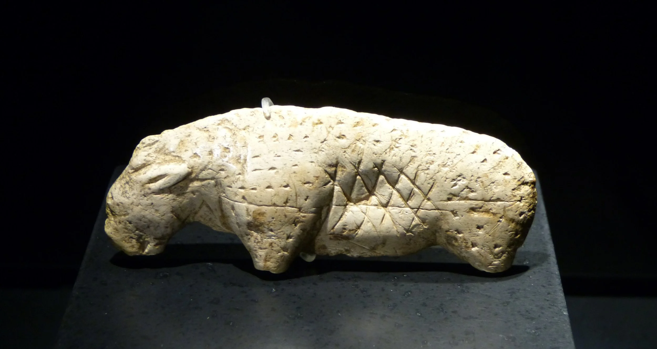 A two-inch long, abstract figure, deemed a lion, made from ivory, incised with two rows of diamond patterns made from cross-hatching, and covered with small, triangular impressions. c. 33,000 BCE. Vogelherd cave, Germany