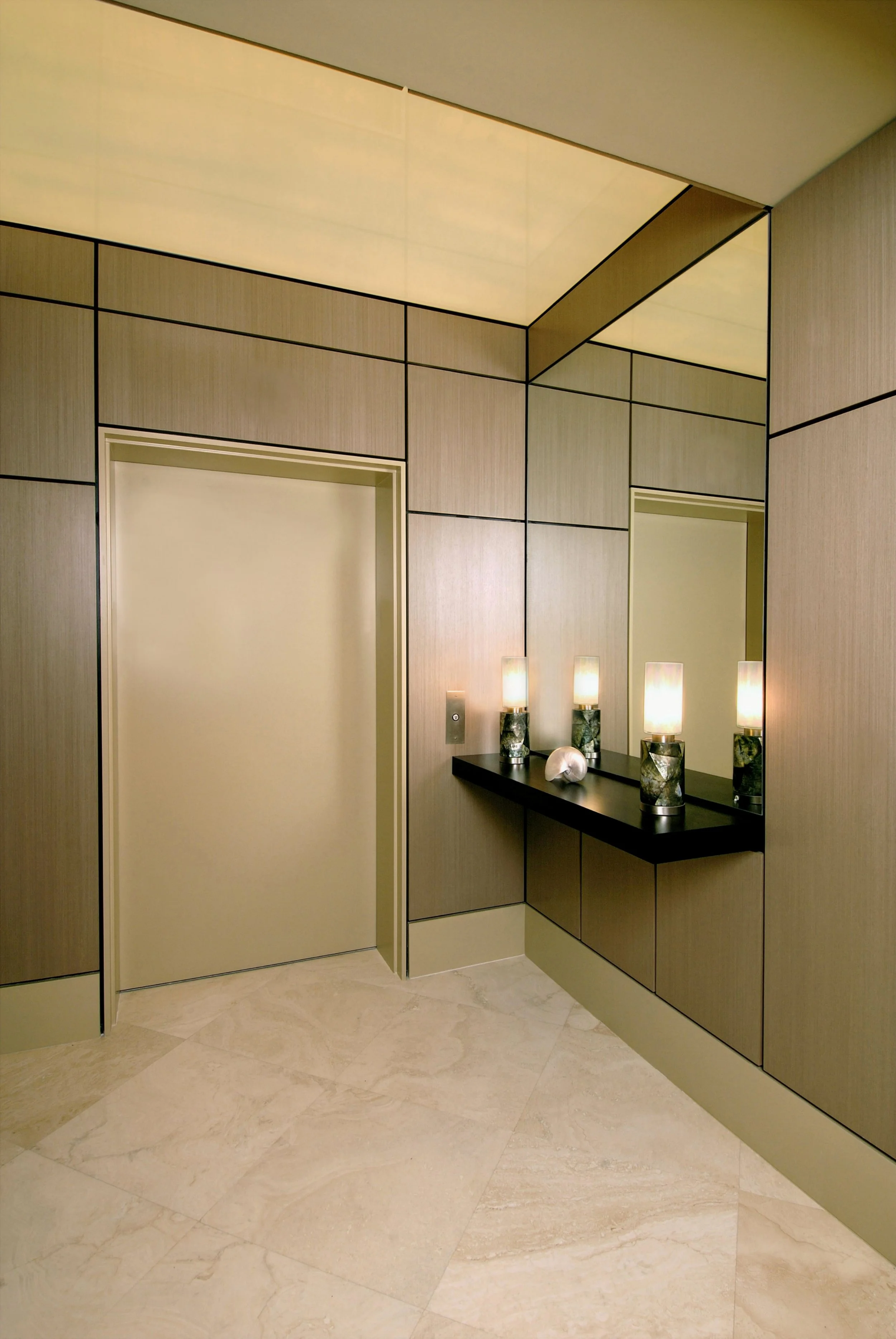 Small, crisply designed elevator lobby with wood paneling, mirror, luminous ceiling, floating shelf with pair of black mother of pearl lamps