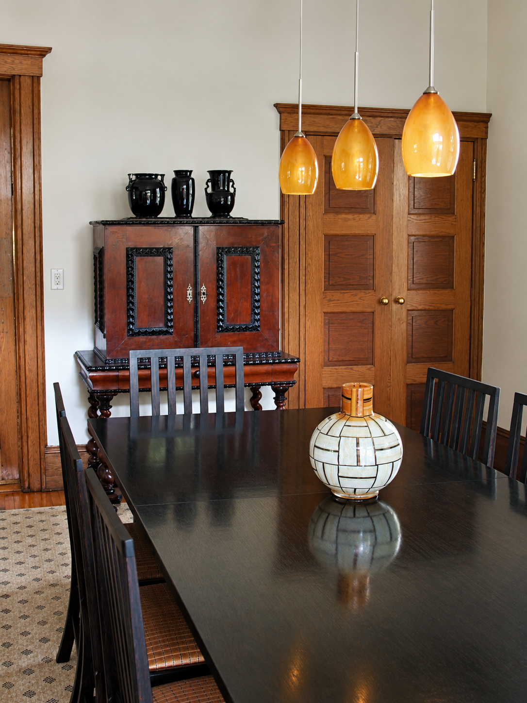 a dining room wit th partial view of table and chairs, a major glass centerpiece, 3 amber glass pendant fixtures, a 17th century cabinet on a stand, with 3 black vases on top