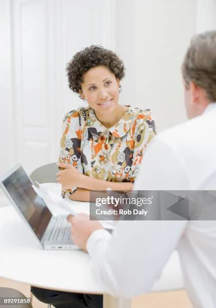 A woman and a man having a conversation at a table in a bright, white room. The woman is smiling and has short, curly hair, wearing a colorful, patterned blouse. The man is partially visible, wearing a white shirt, and holding a tablet. There is a laptop open on the table.