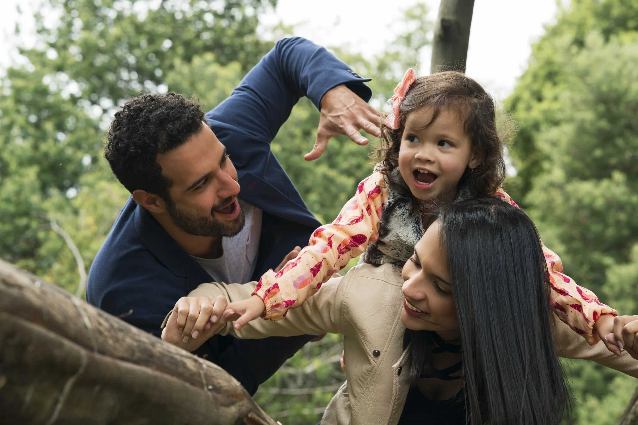 Family outdoors during daytime, with a man, woman, and two young girls playing and climbing on a tree, surrounded by green trees.