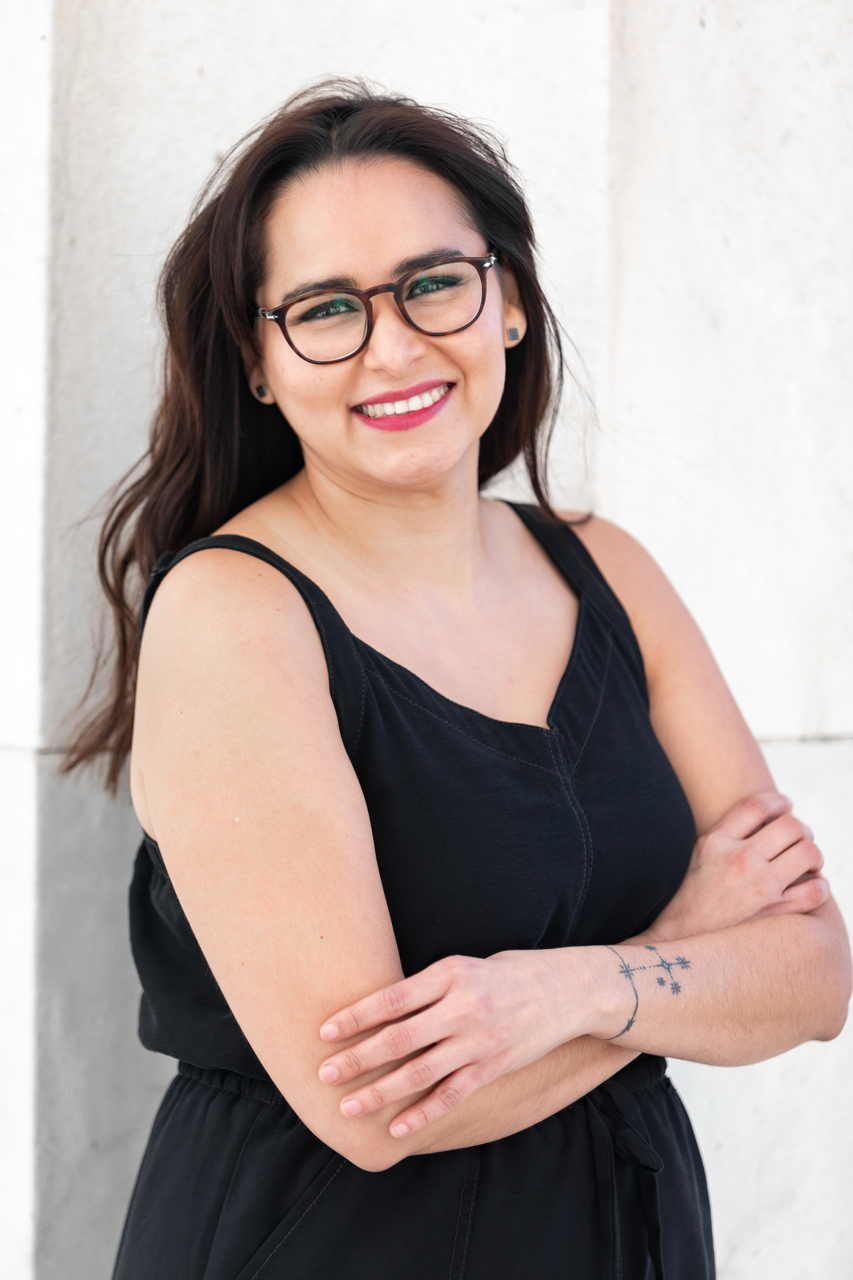 A woman with glasses and long dark hair smiling and crossing her arms, standing against a light-colored wall.
