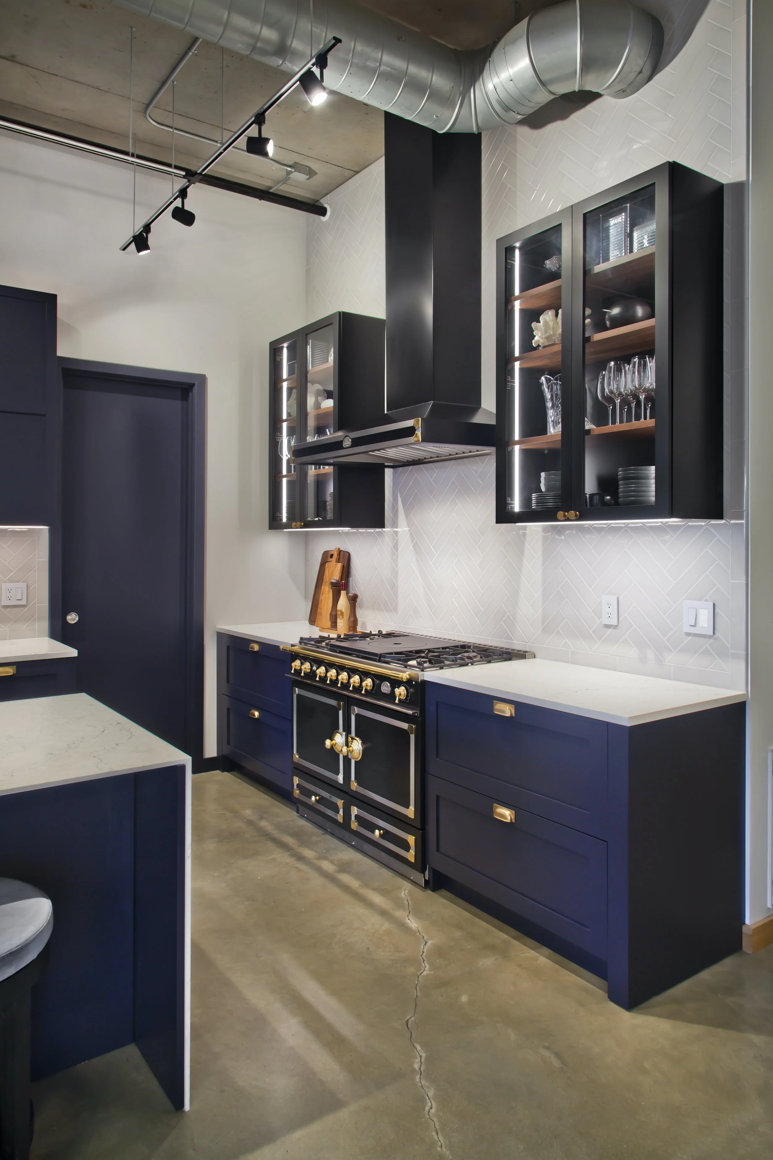 Modern kitchen with black cabinetry, white countertops, and a black oven with gold accents. Open glass-front cabinets display glassware and dishes. Exposed concrete floor with a crack, industrial ceiling with ductwork, and track lighting.