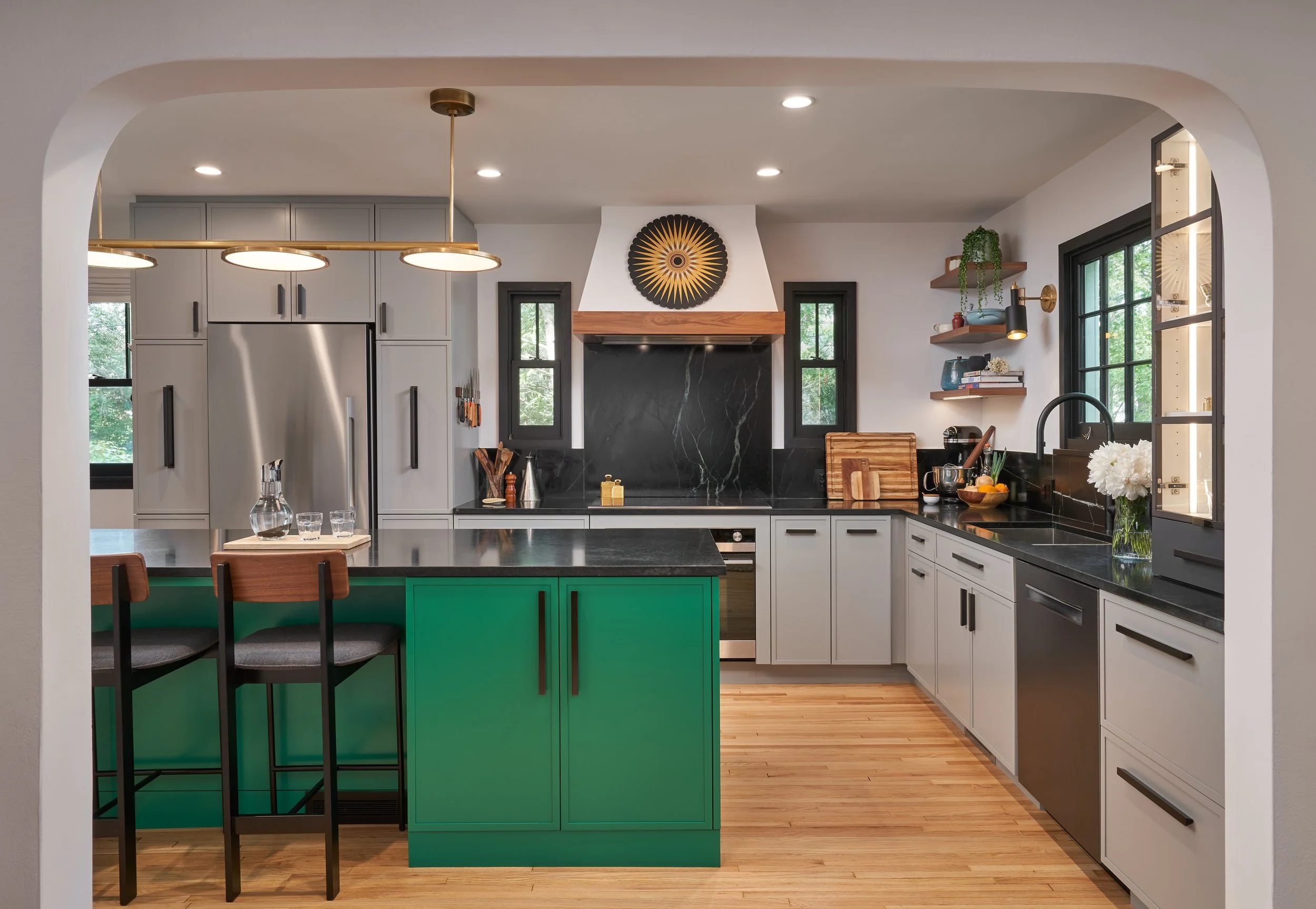 Modern kitchen with gray cabinets, black countertops, and a green island. Features a stainless steel refrigerator, open wooden shelving, and black framed windows, with hardwood floors.