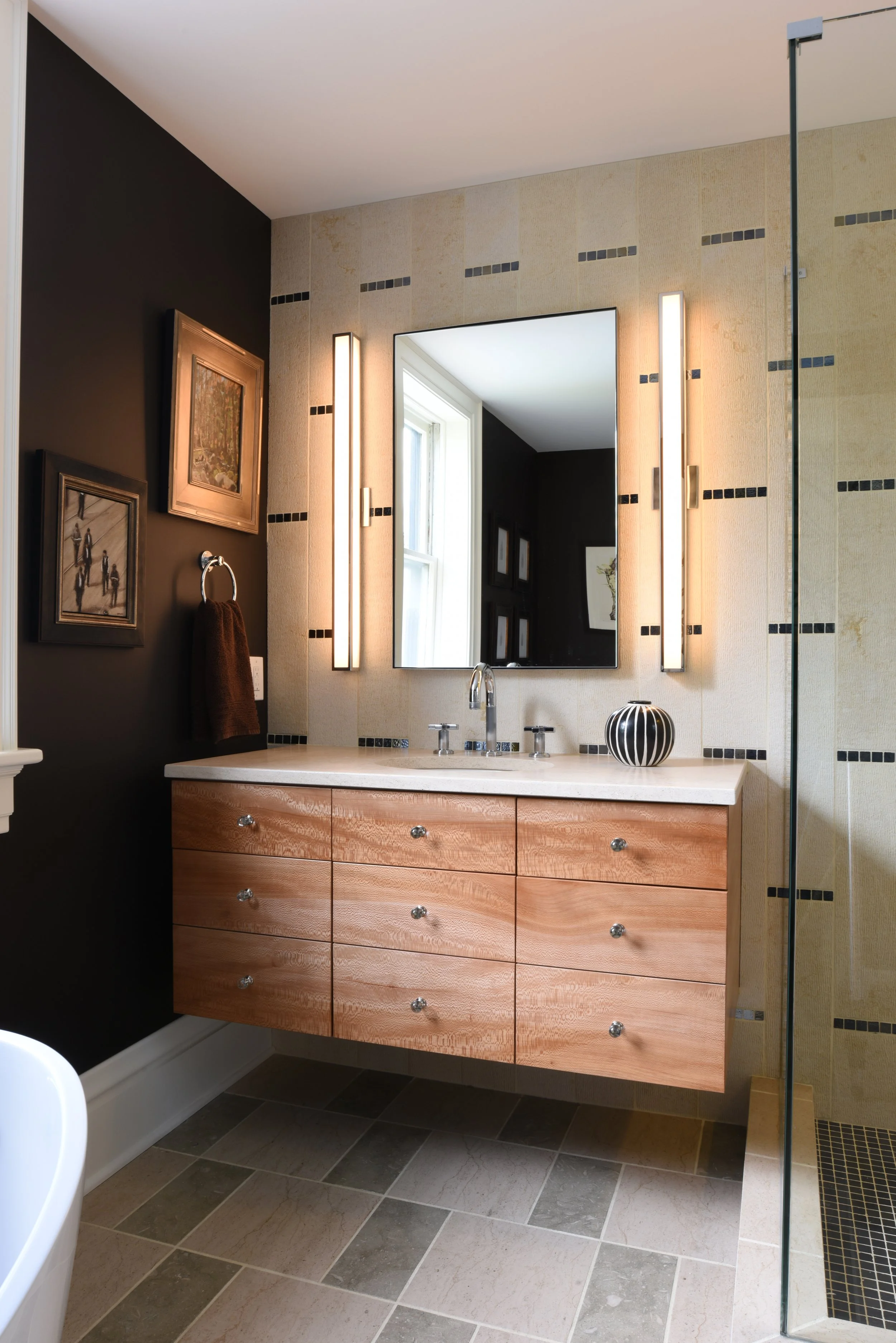 Modern bathroom with black and beige tile walls, wooden vanity with six drawers, large mirror, sleek vertical lighting, and a shower with glass partition.