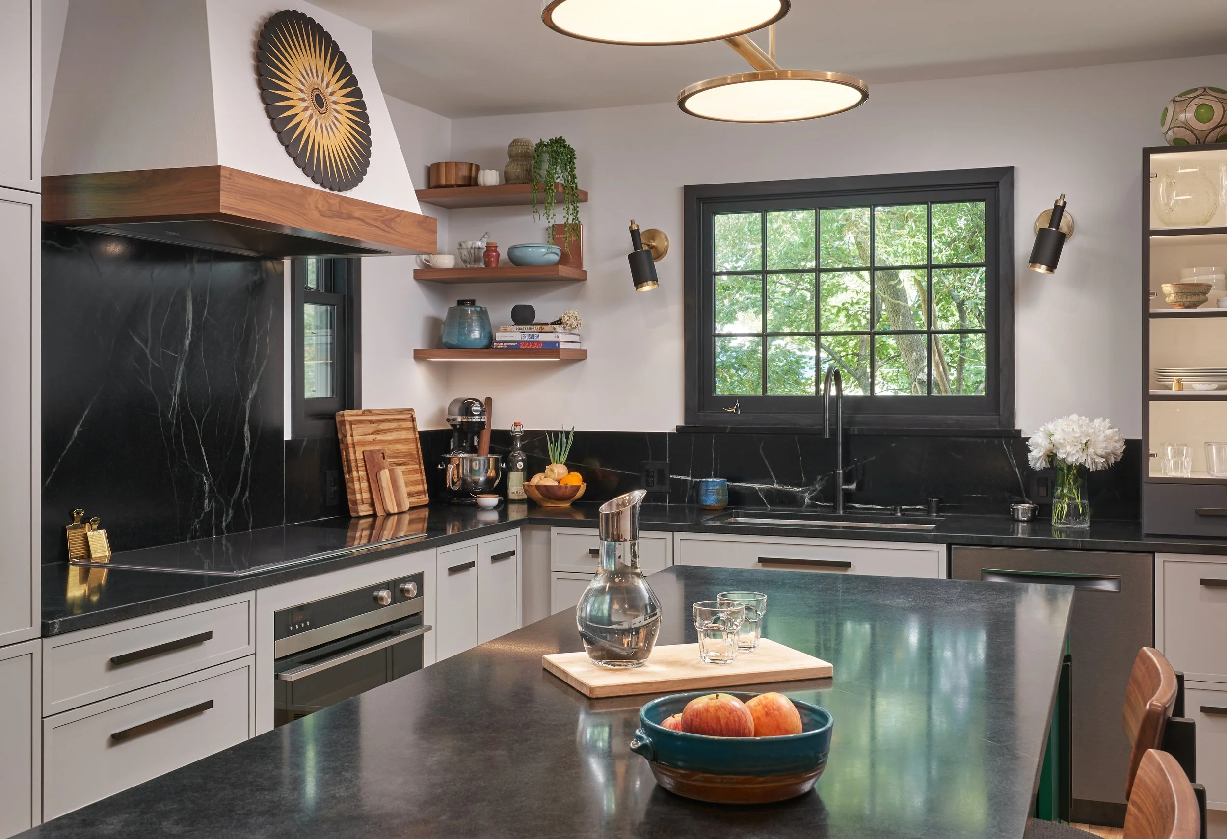 Modern kitchen with white cabinets, black marble countertops, open shelving, and a large black-framed window showing green trees outside.