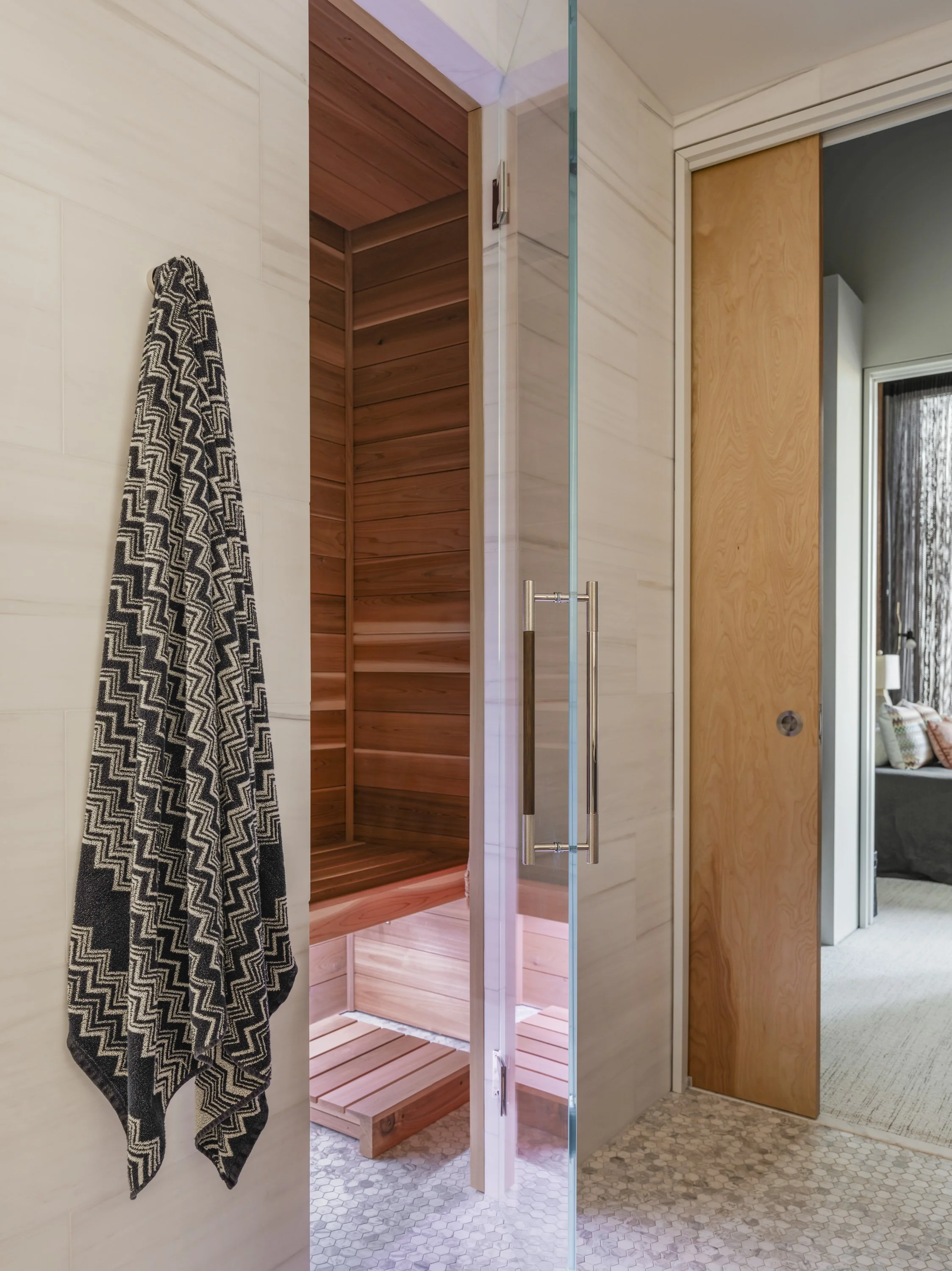 A bathroom with a glass door, a black-and-white chevron towel hanging on the wall, a wooden door to a sauna, and a doorway to a living room.