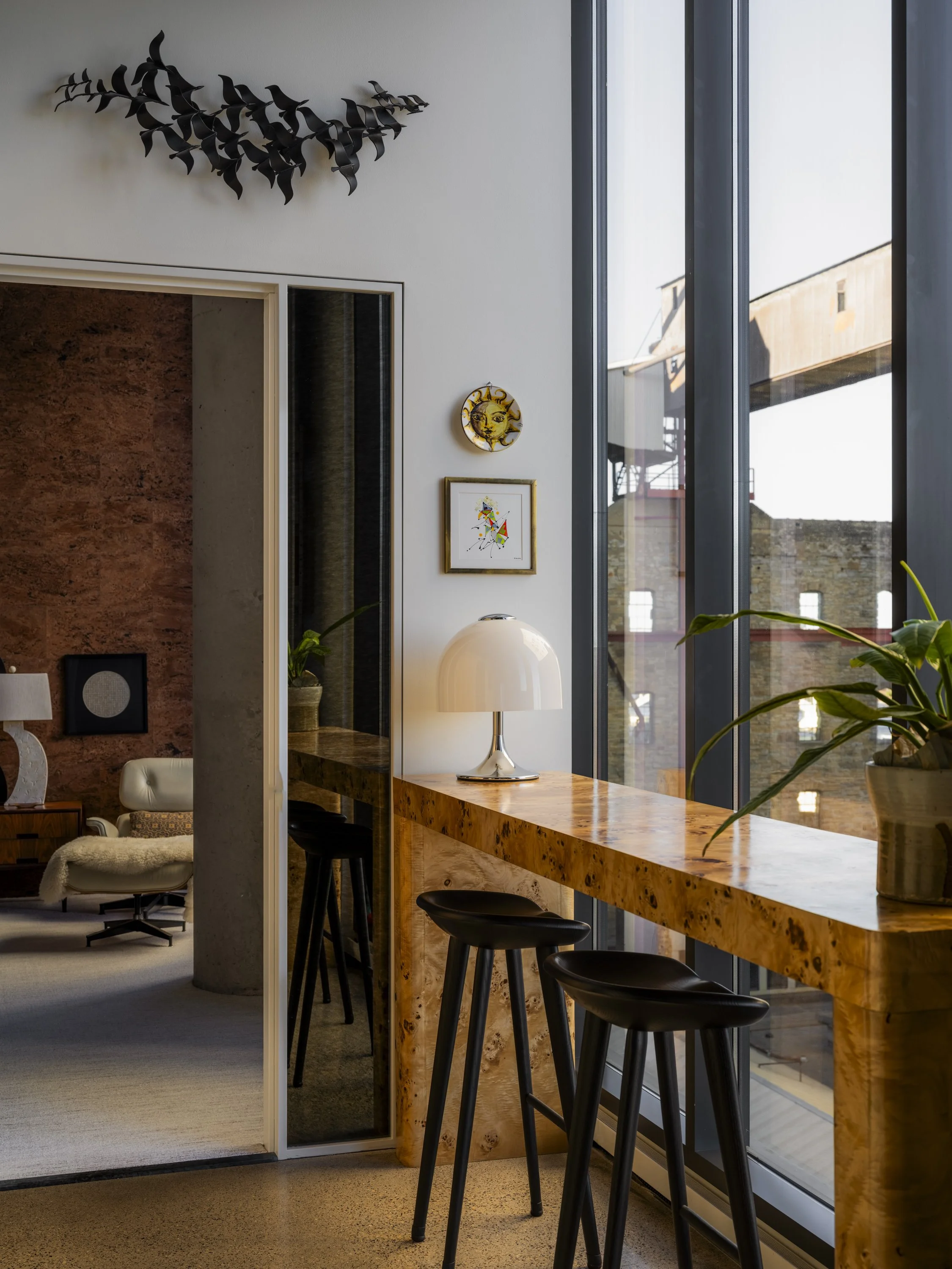 Interior of a modern apartment with a high wooden counter, black bar stools, a white table lamp, a potted plant, and artwork on the wall near large windows