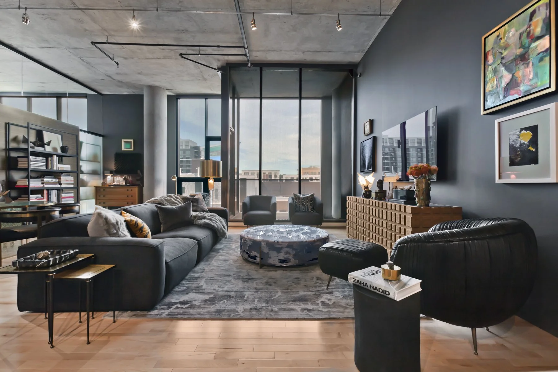 Modern living room with large windows, gray walls, and contemporary furniture including a black sofa, armchairs, and a round ottoman. Decor includes framed art and a wooden media console with a mounted TV.