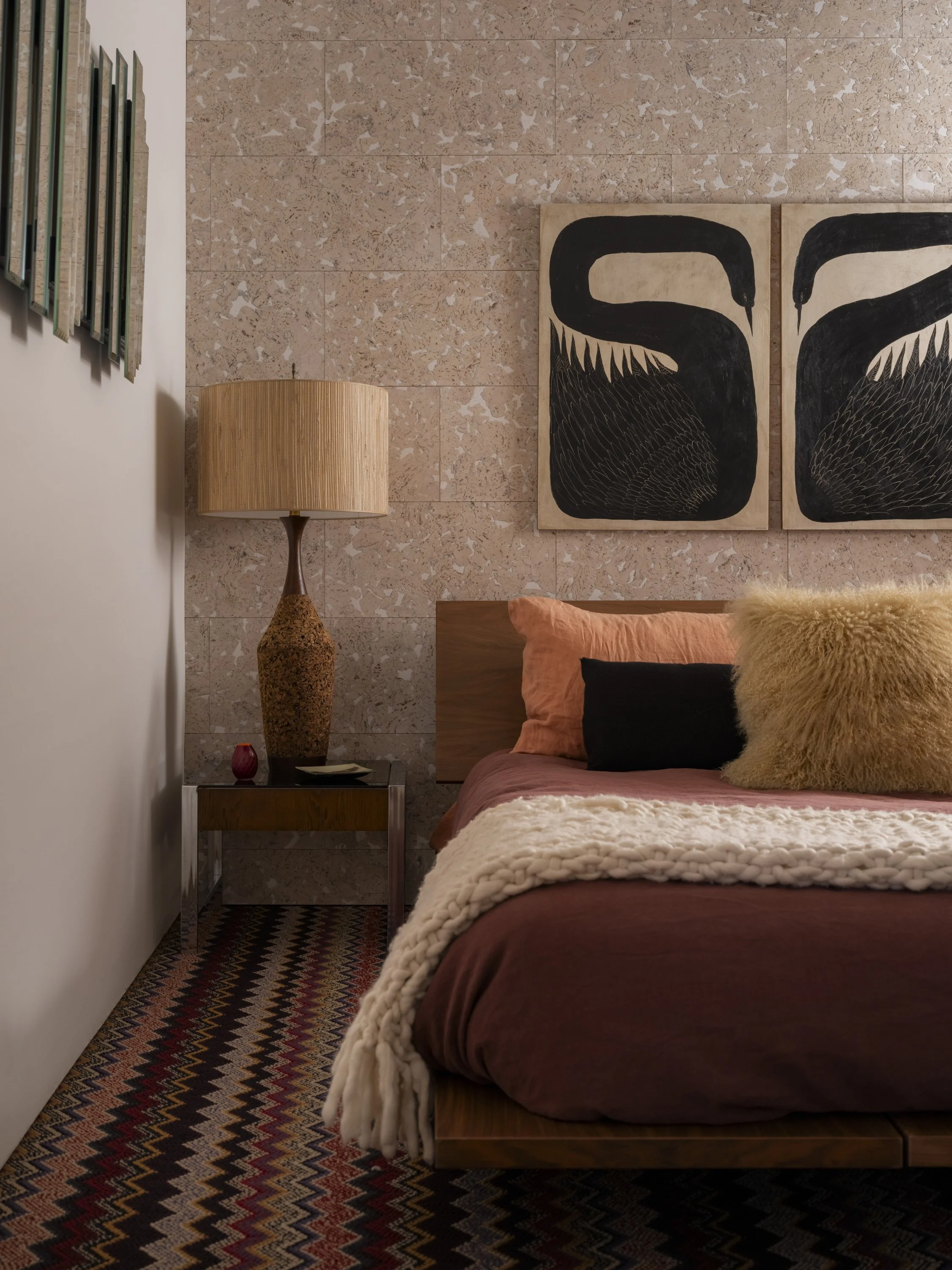 A cozy bedroom with a wooden bed frame, rust-colored bedding, and pillows, including a large fluffy beige pillow. Beside the bed is a wooden nightstand with a tall, textured lamp, a small red decorative item, and a plate. The wall behind the bed feat