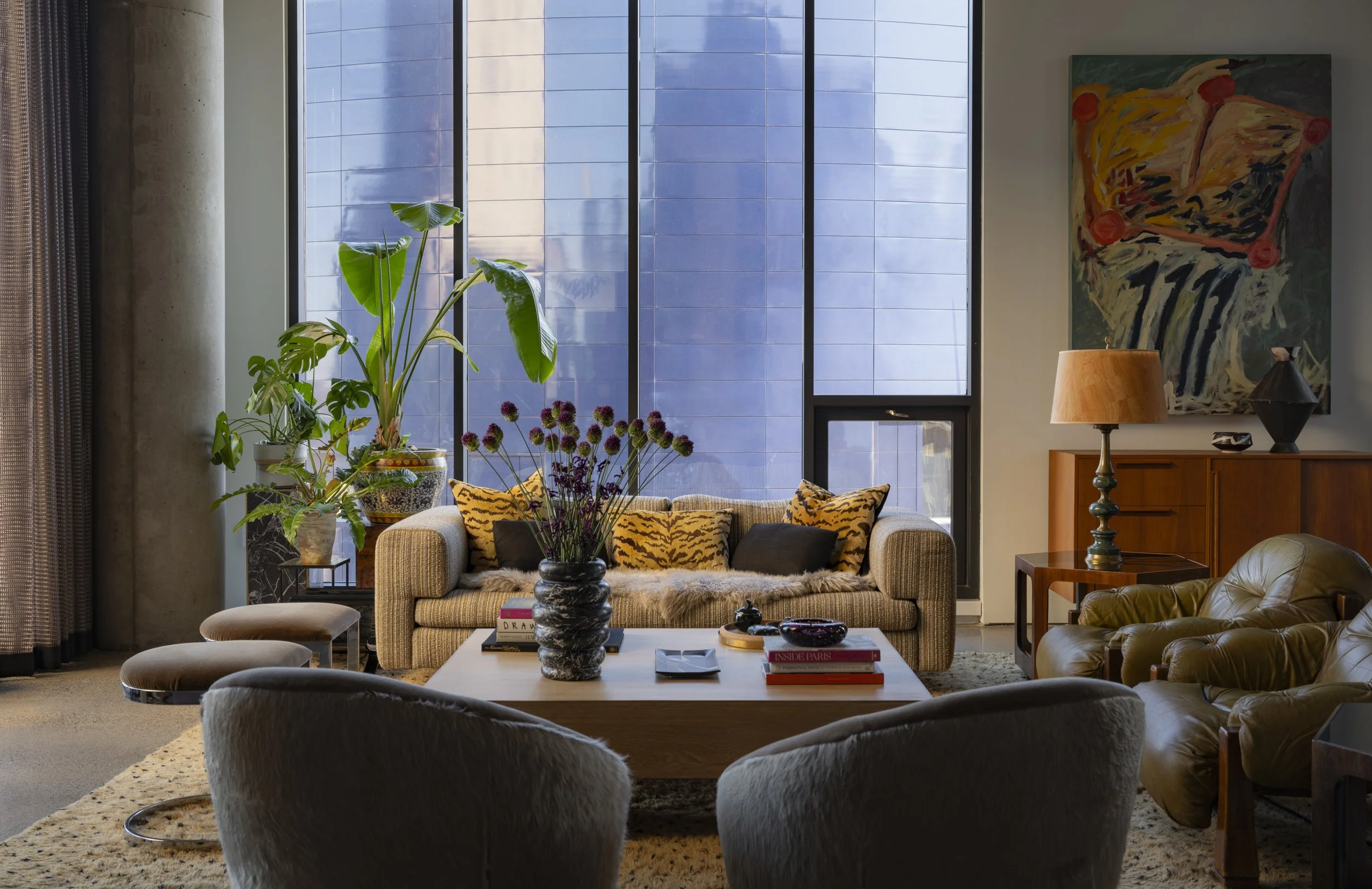 Living room with large windows, a beige sofa with tiger-patterned and dark cushions, a coffee table with books and a flower vase, a leather armchair, a side table with a lamp, an abstract painting on the wall, and houseplants.