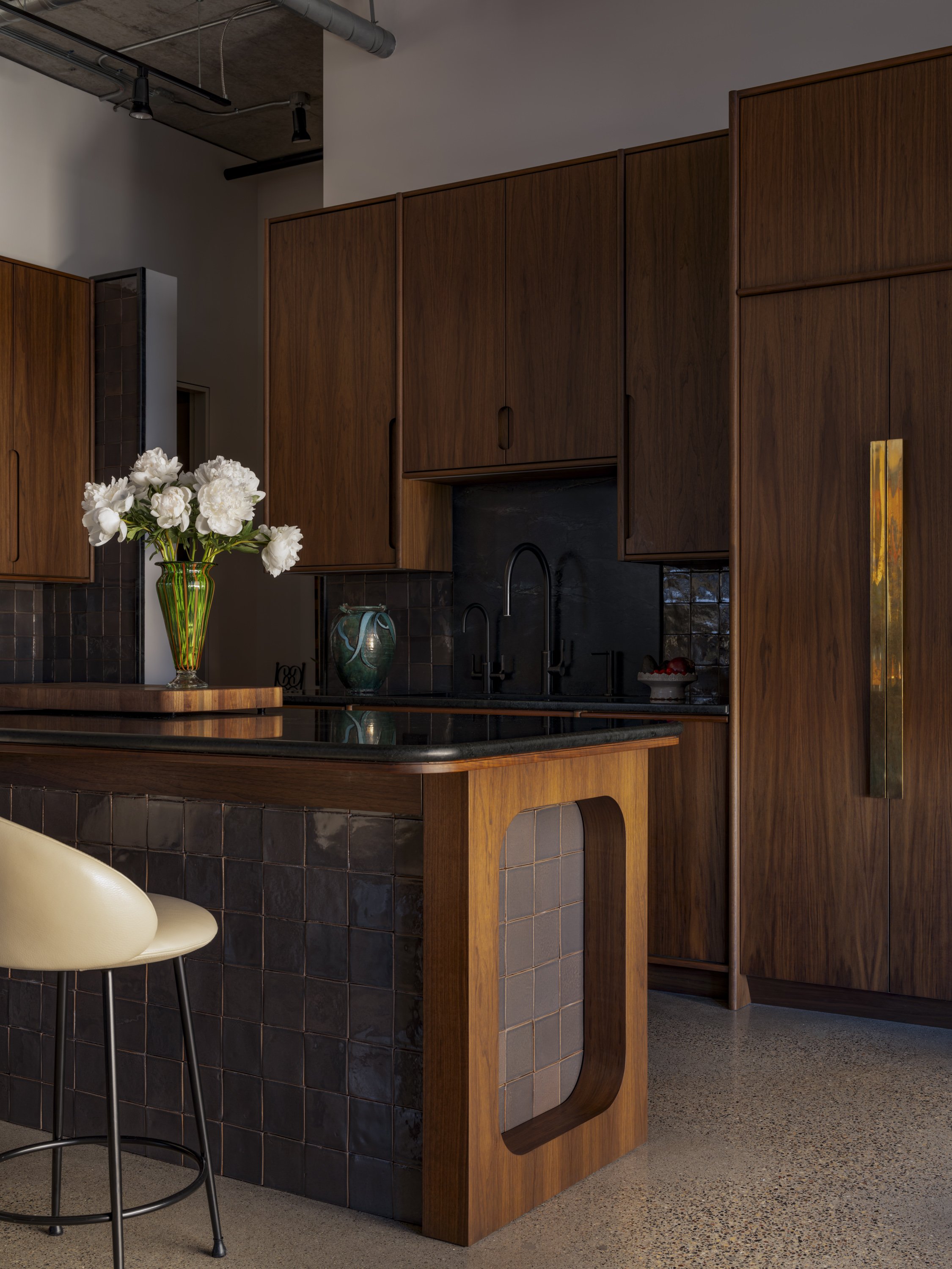 A modern kitchen with dark wood cabinets, a black countertop, a vase of white flowers, and a black tiled backsplash. There is a barstool with a cream-colored seat in the foreground.