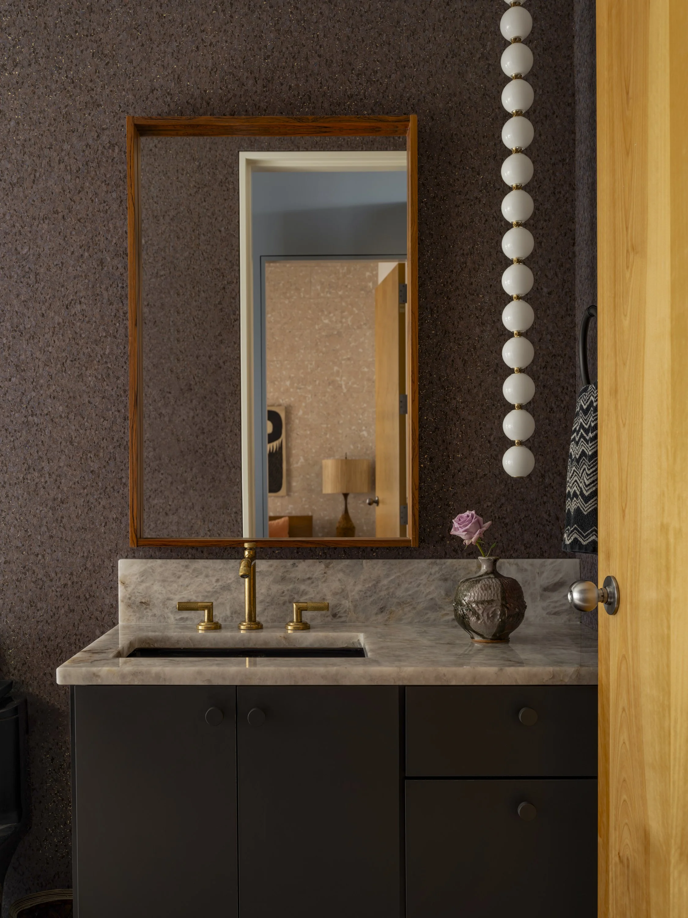 A bathroom vanity with a marble countertop, a faucet, and a black cabinet with drawers, a vase with a purple flower, a large rectangular mirror, dark textured walls, a string of white globe lights, and a wooden door slightly ajar.
