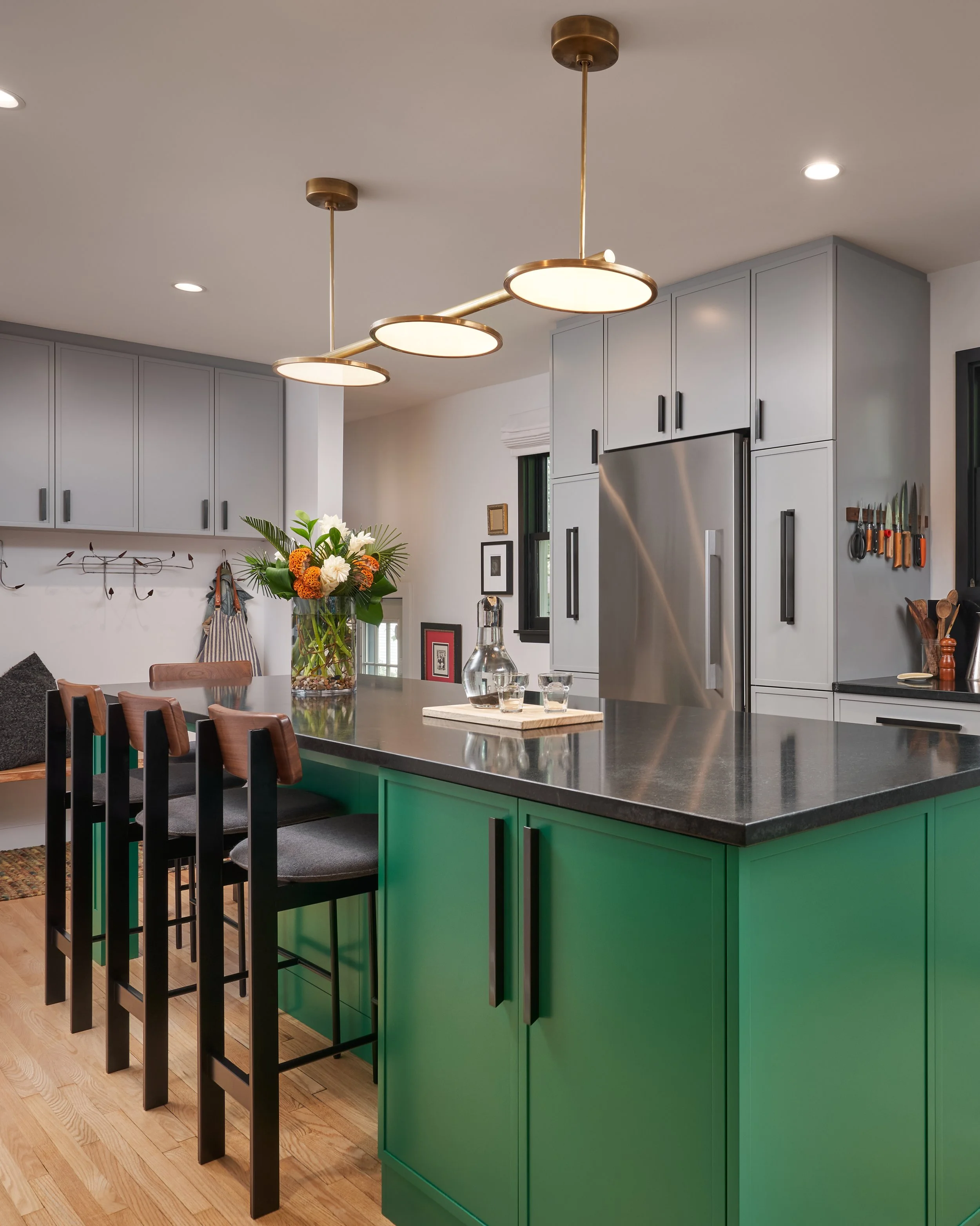 Modern kitchen with green island, gray cabinets, black handles, and gold pendant lights. Decor includes a flower vase, glassware, and a knife rack.
