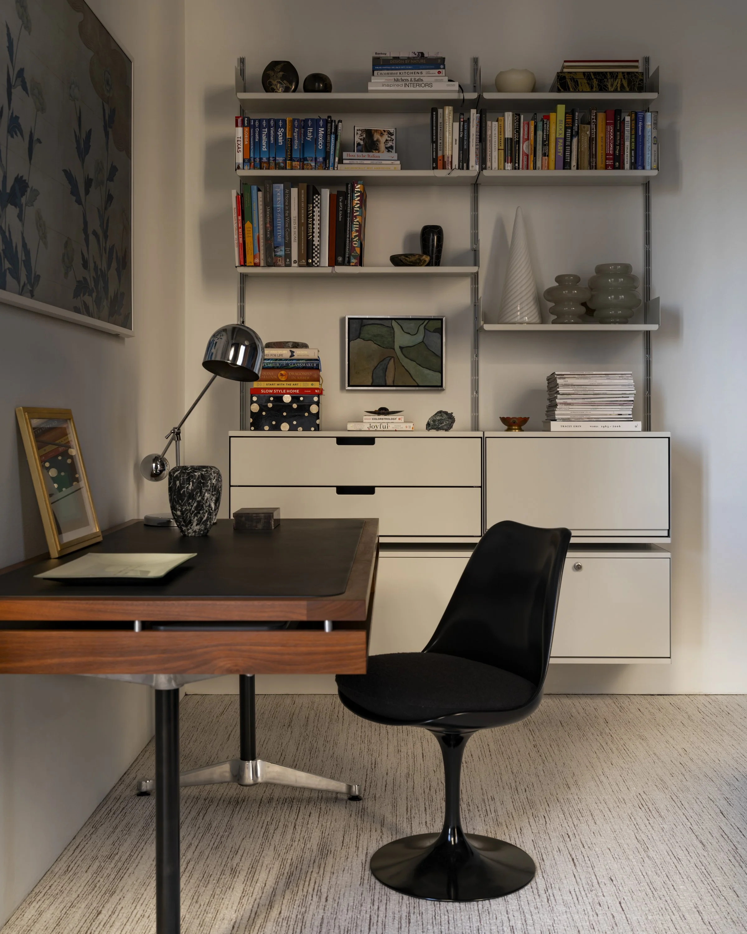 A modern home office with a wooden desk, black chair, white storage cabinet, and a wall-mounted bookshelf filled with books and decorative items.